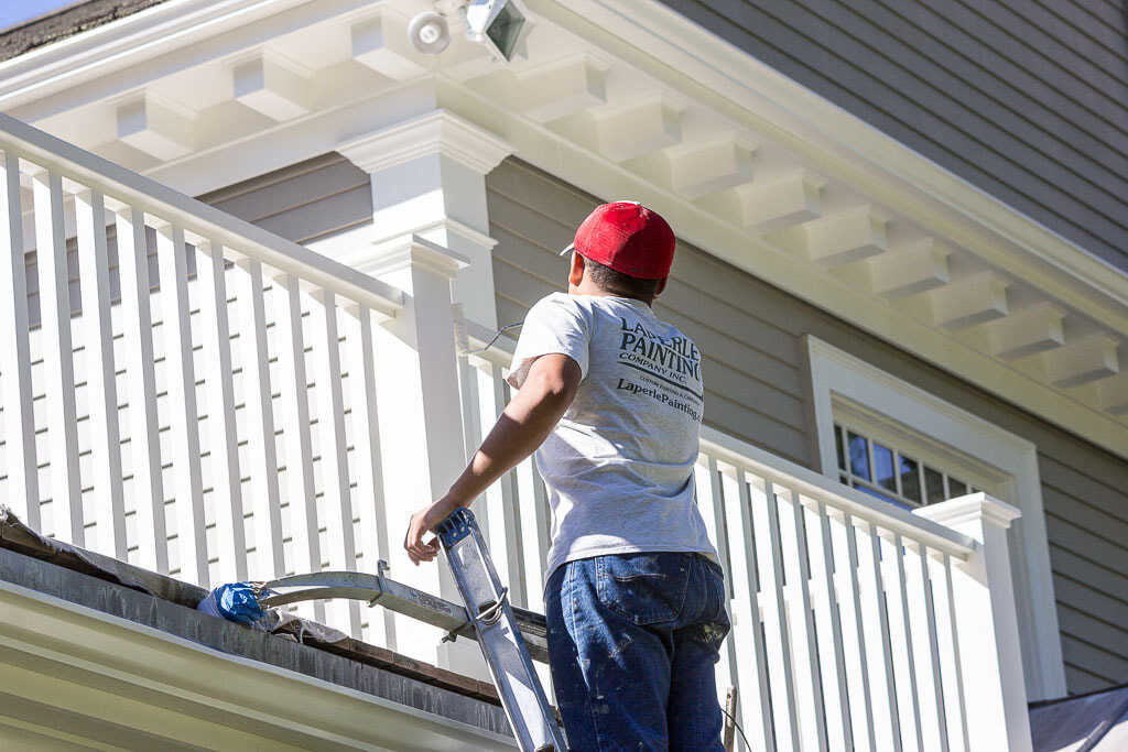 Painting Contractor in Dover, MA Since 1992 Person in a red cap painting a house’s white exterior while standing on a ladder