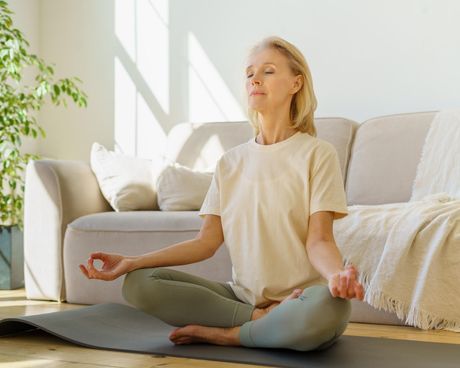 A woman is sitting in a lotus position on a yoga mat in a living room.