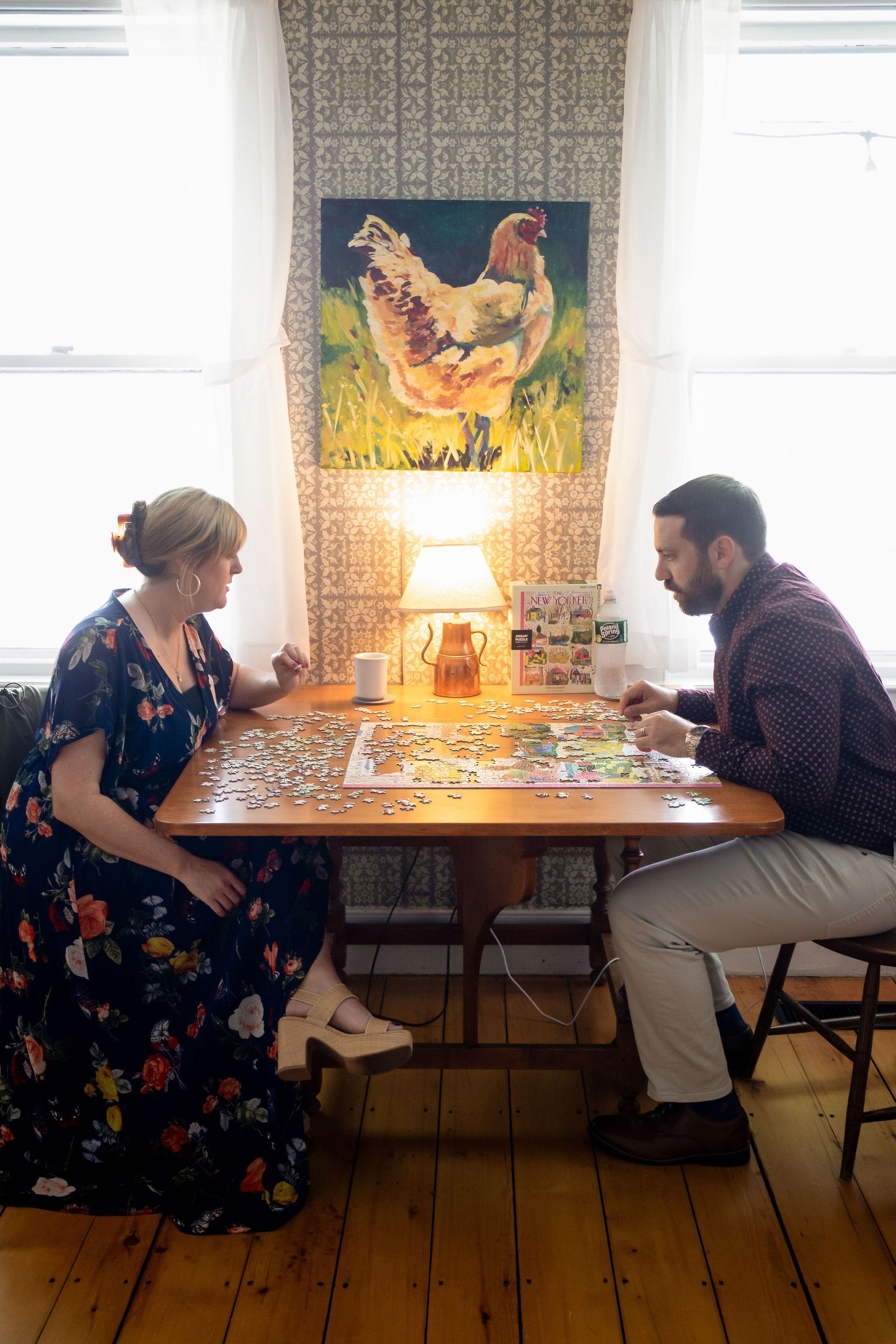 Woman and man sitting at a table working on a jigsaw puzzle, lit by a lamp, with chicken art in the background.