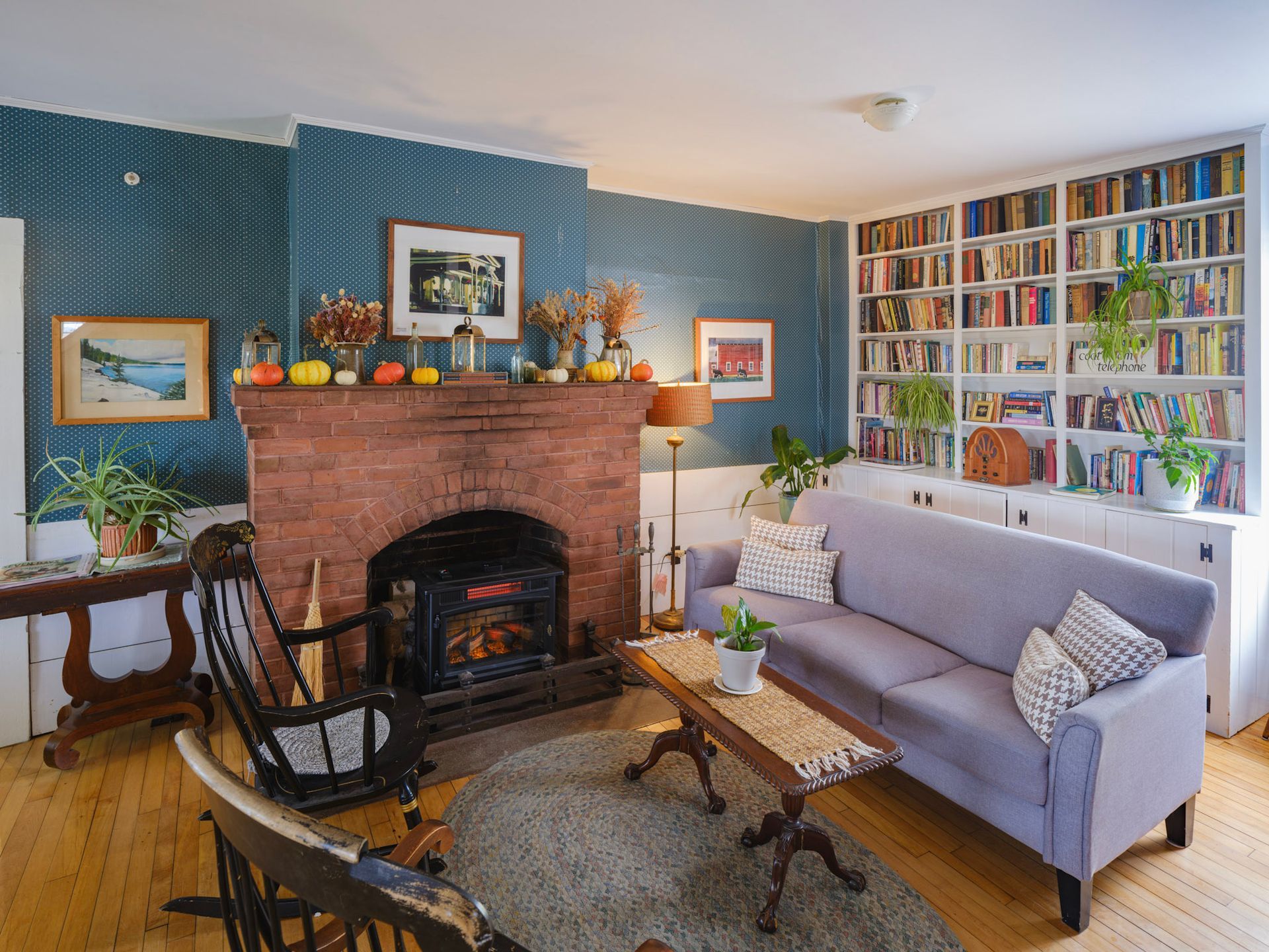 Cozy living room with brick fireplace, bookshelves, light blue sofa, wooden floors, and patterned blue walls.