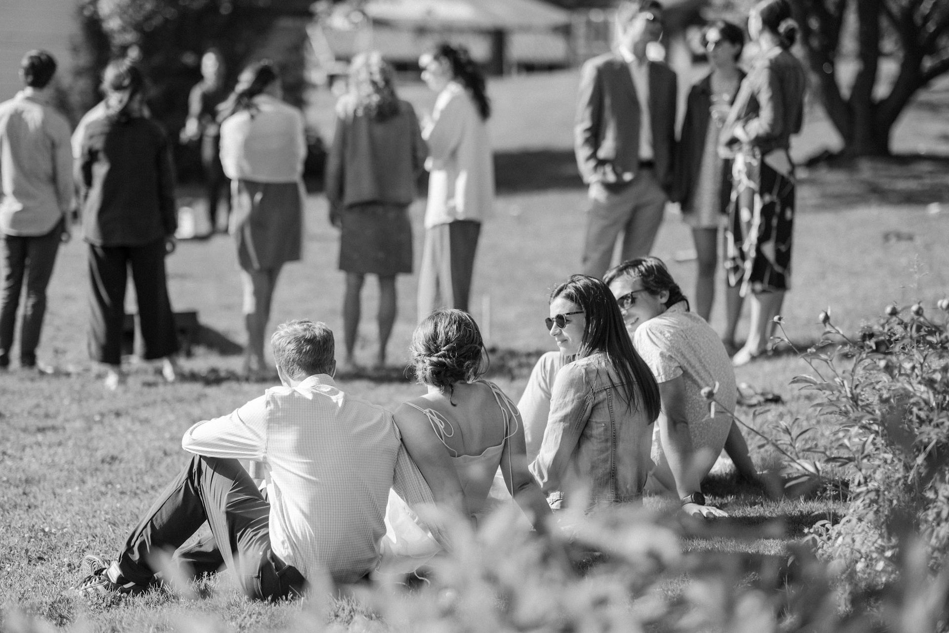 People sitting on grass, looking towards a group standing in the background. Black and white photo.