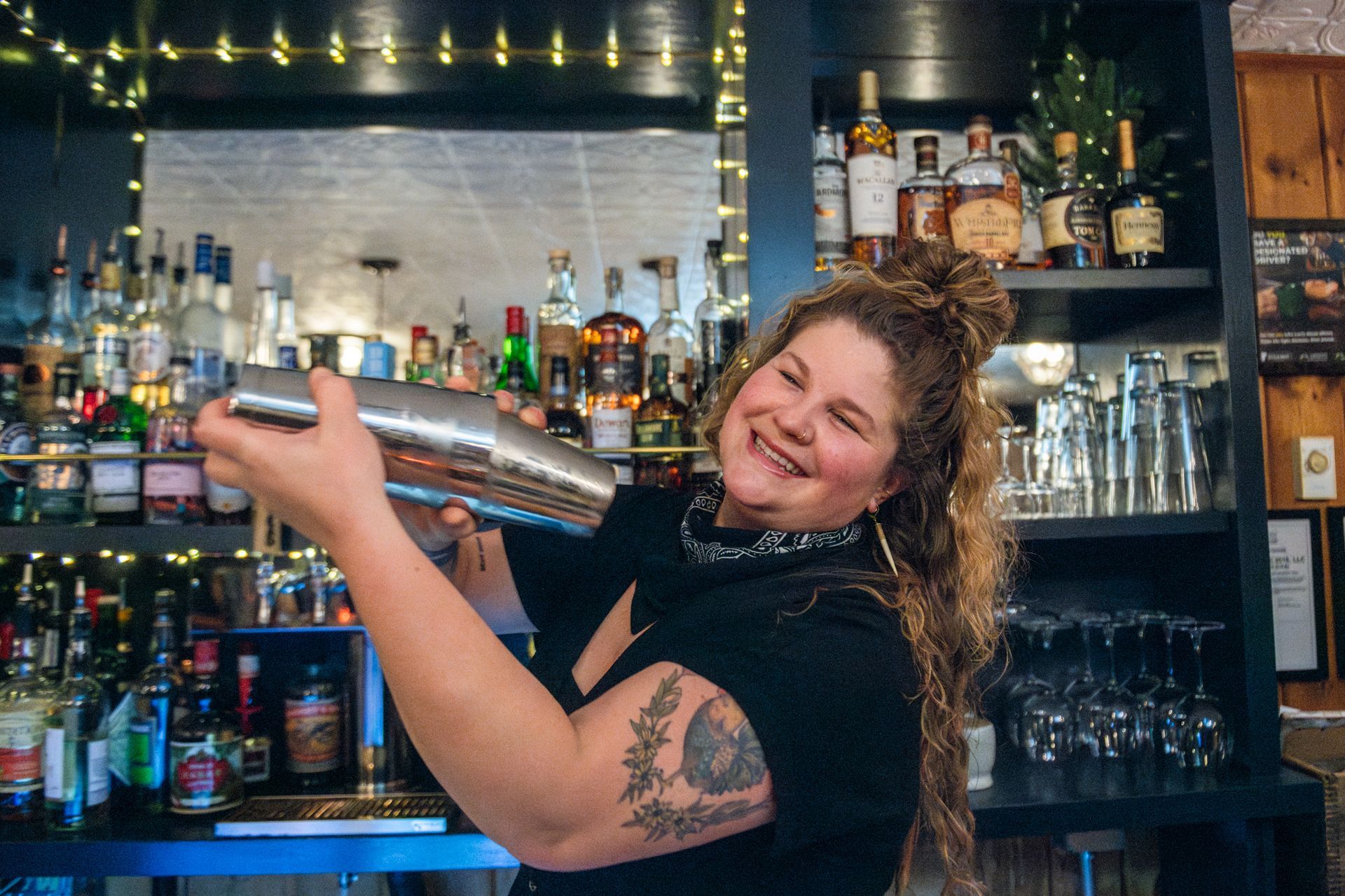 Bartender smiling, shaking cocktail shaker behind bar with bottles.