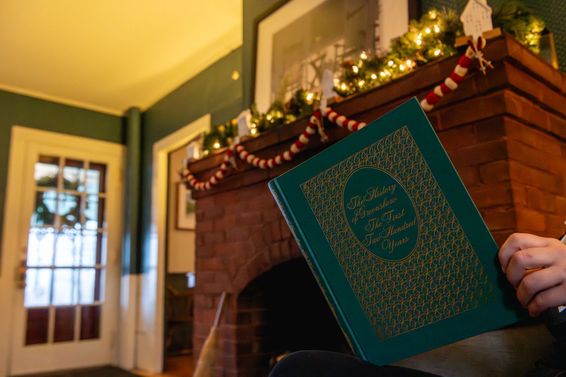 Person holding a green book in front of a fireplace decorated with garland.