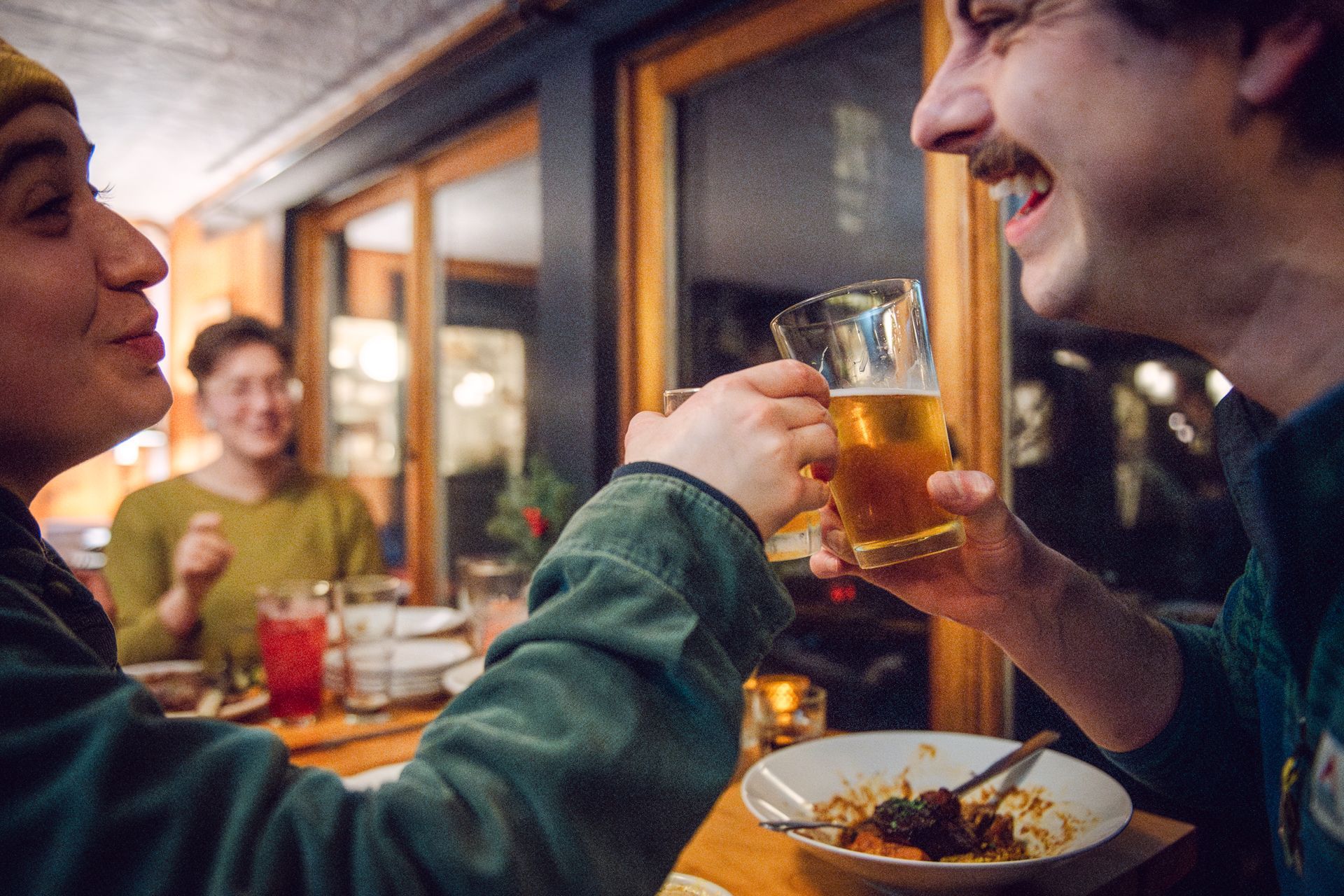 Two people clink beer glasses, smiling. A third person smiles in the background, seated at a table in a restaurant.