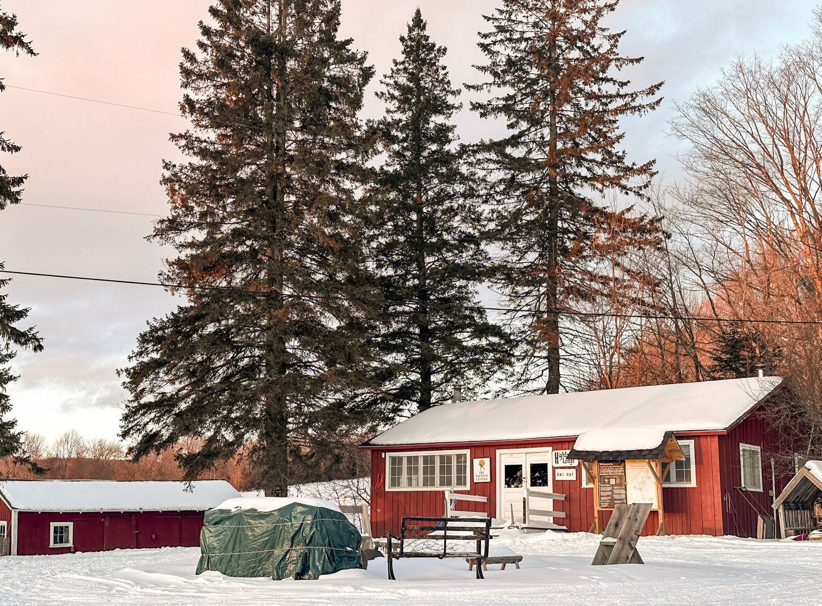Snow-covered red cabins and evergreens, winter landscape. A covered object and a bench in the snow.