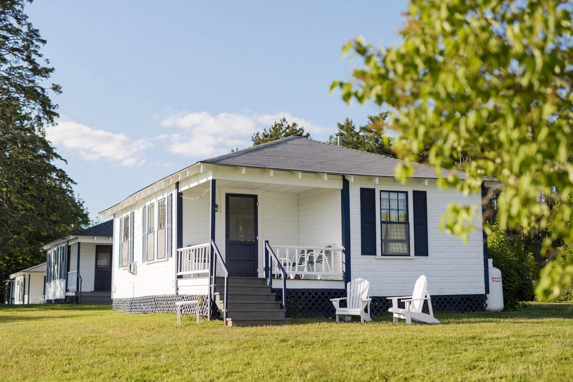 White cottages with blue trim and a grassy yard under a blue sky, two Adirondack chairs sit on the lawn.