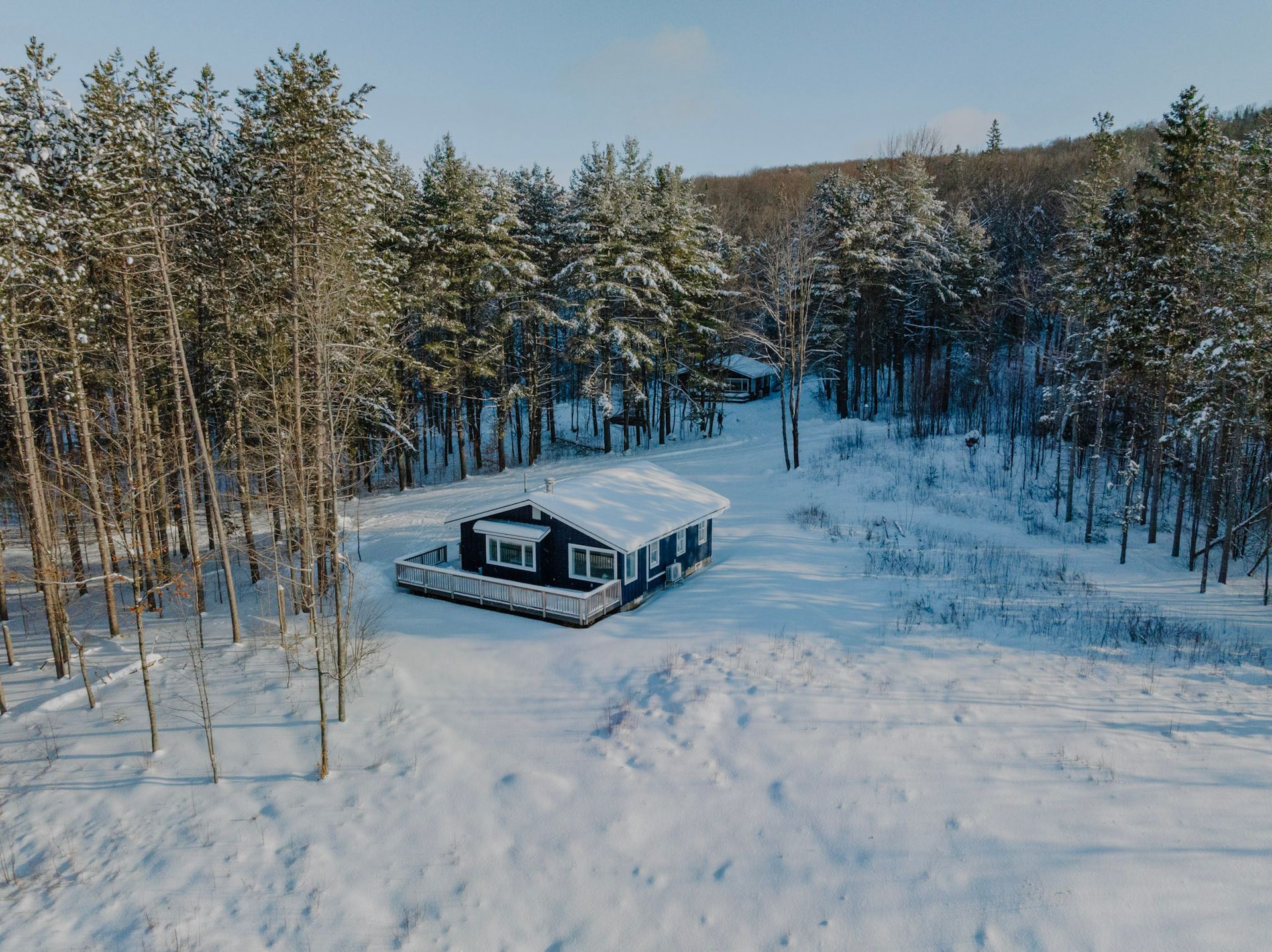 Snow-covered cabin in a forest clearing; trees and a hillside in the background.