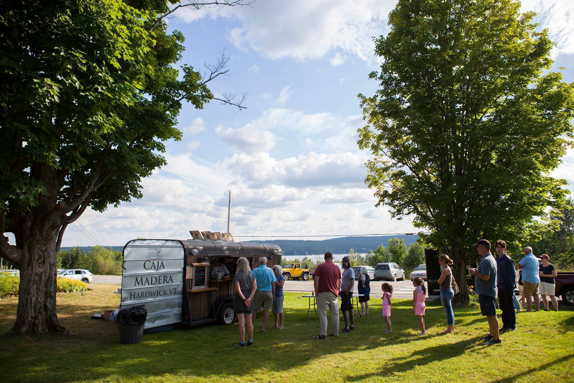 People gathered near a food truck in a park, trees and water visible in the background. Sunny day.