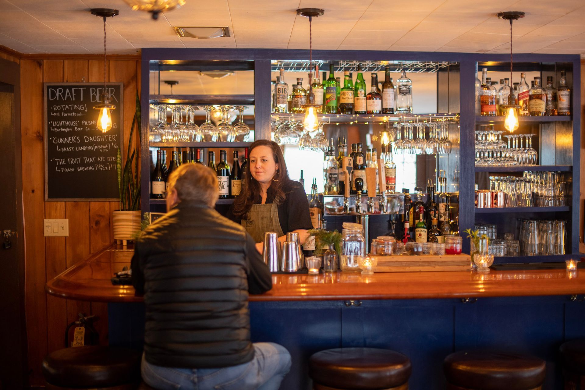 A bartender serves a customer at a cozy bar with liquor bottles on shelves.