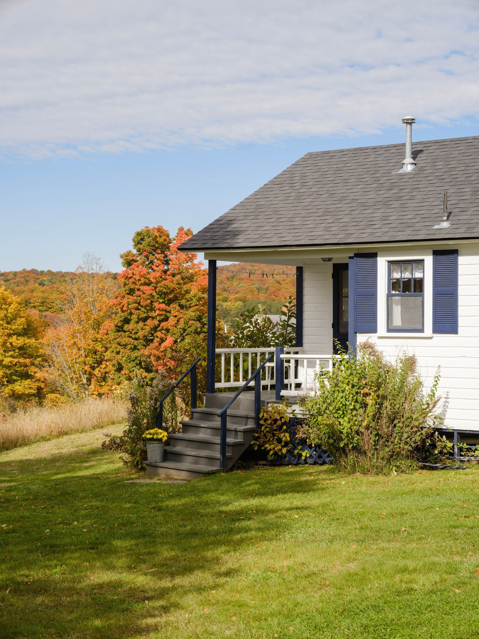 White cottage with dark blue accents and steps; fall foliage in the background.