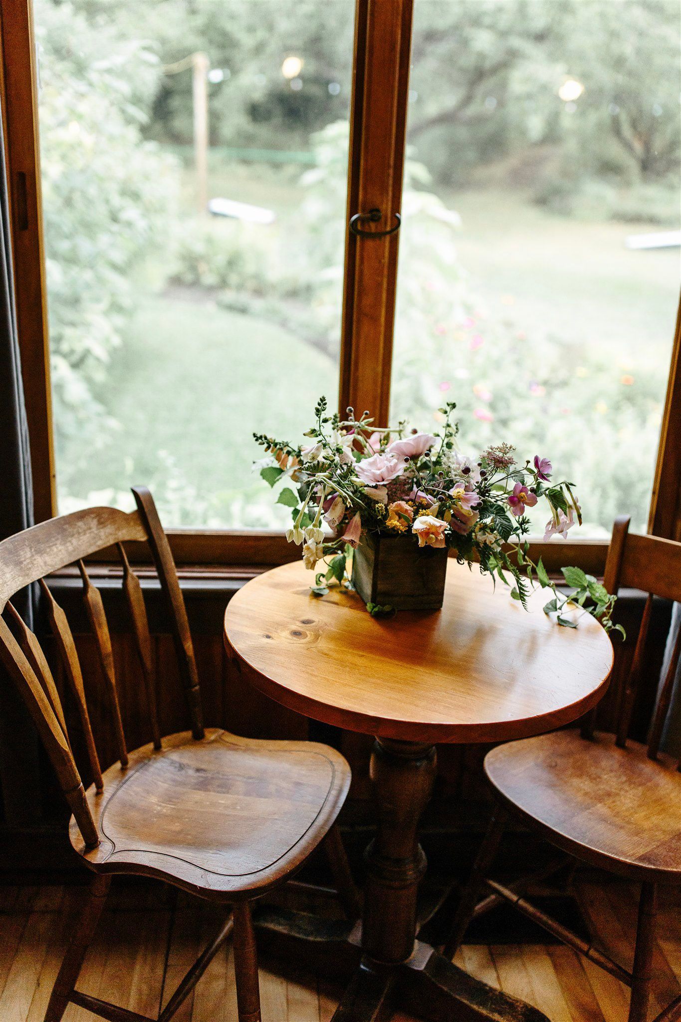 Wooden table with floral arrangement, flanked by two wooden chairs, in front of a window overlooking greenery.