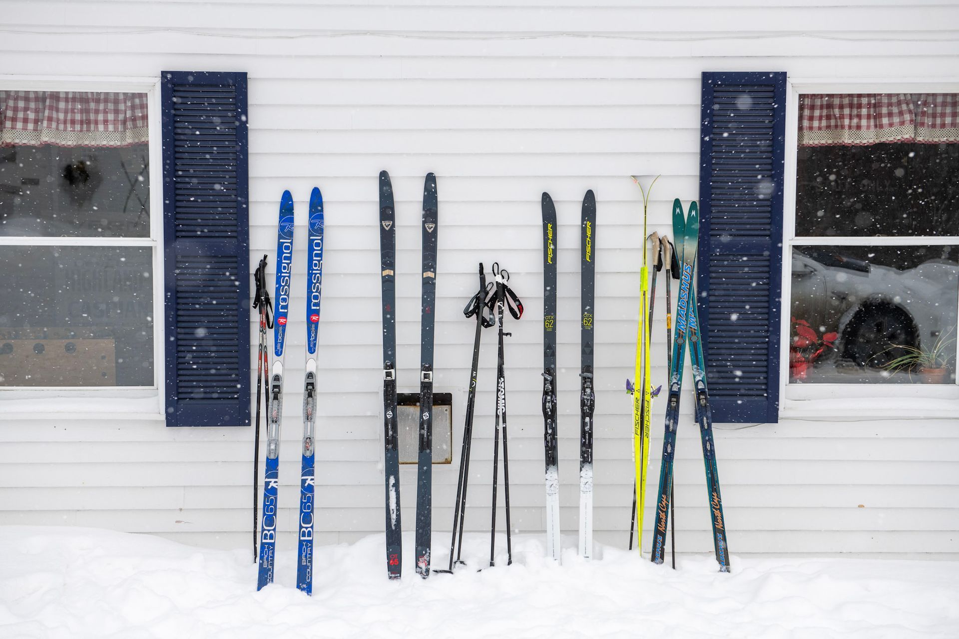 Cross-country skis and poles leaning against a snowy, white building with blue shutters.