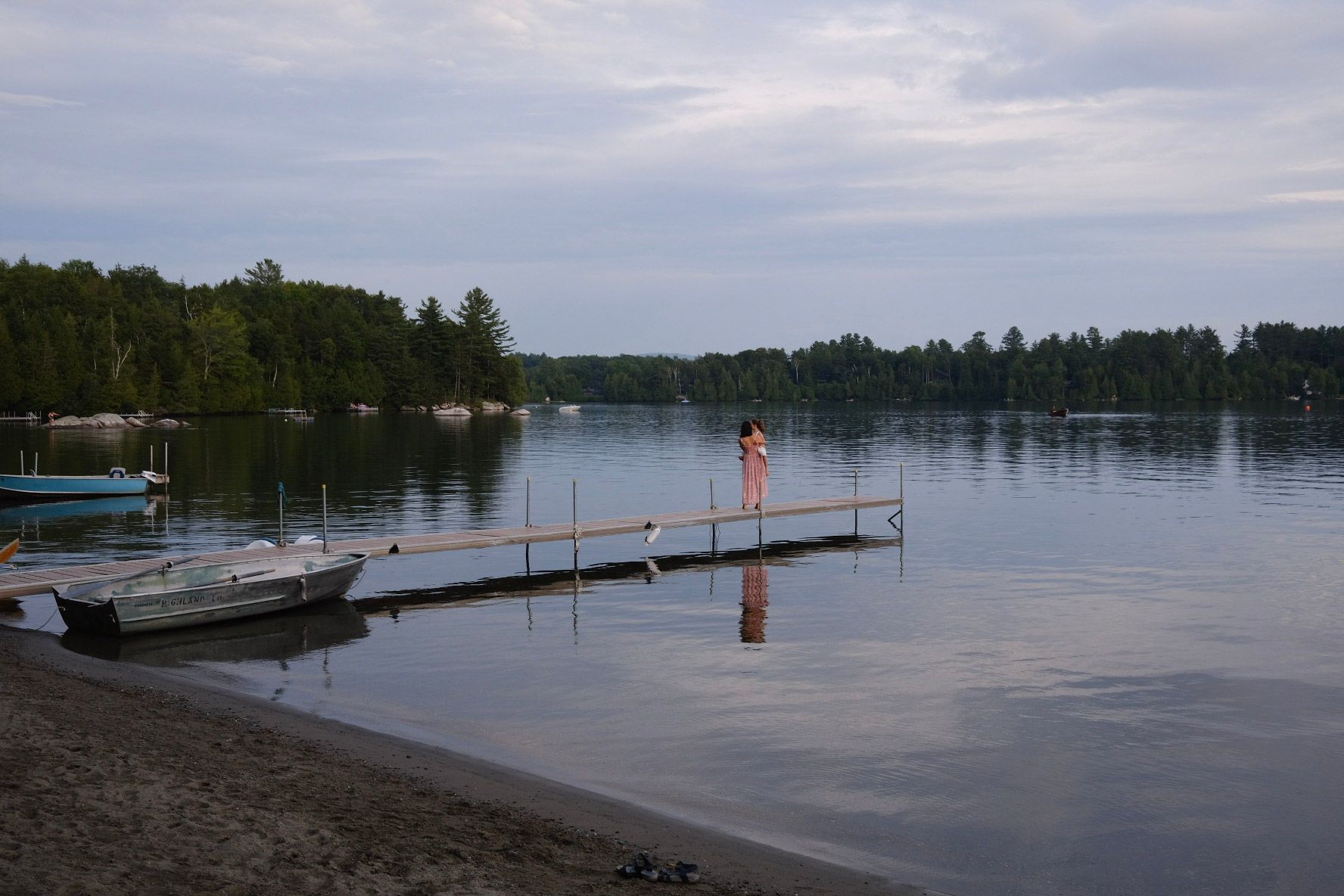 Person standing on a wooden dock over calm water; shore in the foreground, trees in the background.