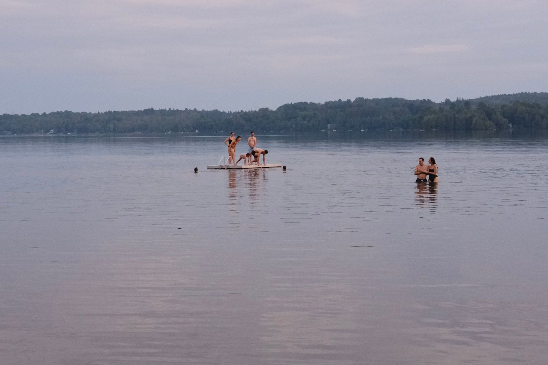 People on a raft and in the water in a lake, trees in the background, overcast sky.