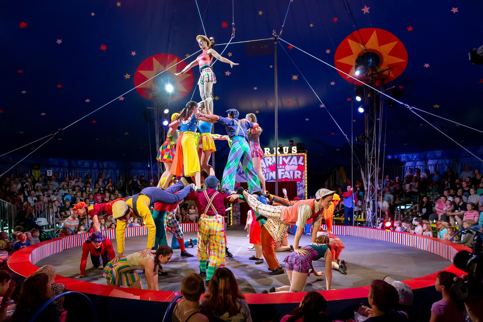 Circus performers balancing in a human pyramid under a big top tent, audience watching.
