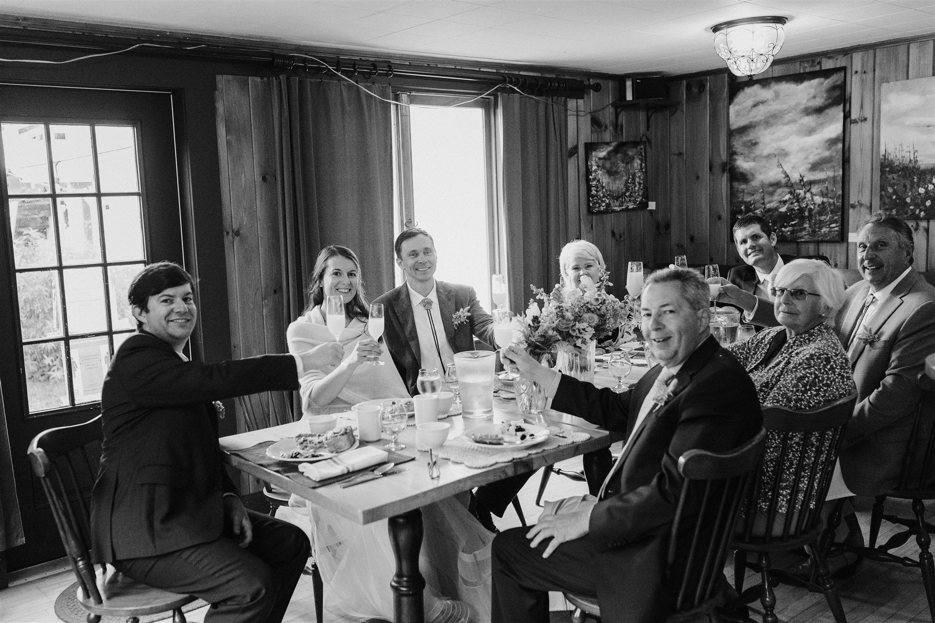 Wedding party seated around a table, toasting. Bride and groom in center, guests smiling in a rustic dining room.