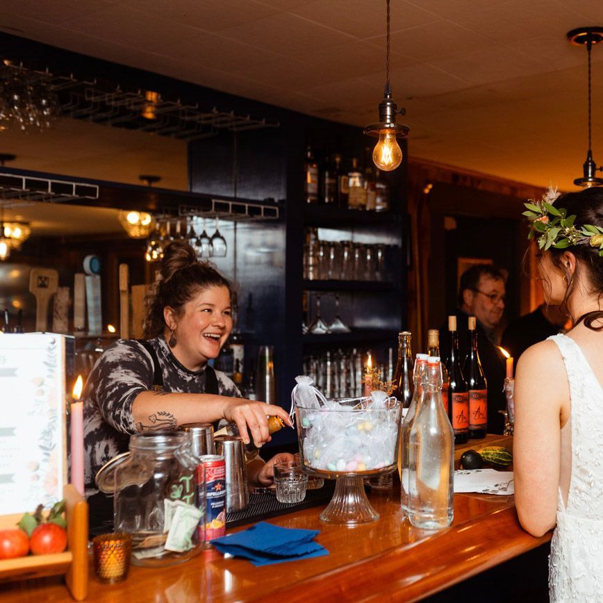 A bartender smiles while pouring a drink. A woman with a flower crown waits at the bar in a cozy setting.