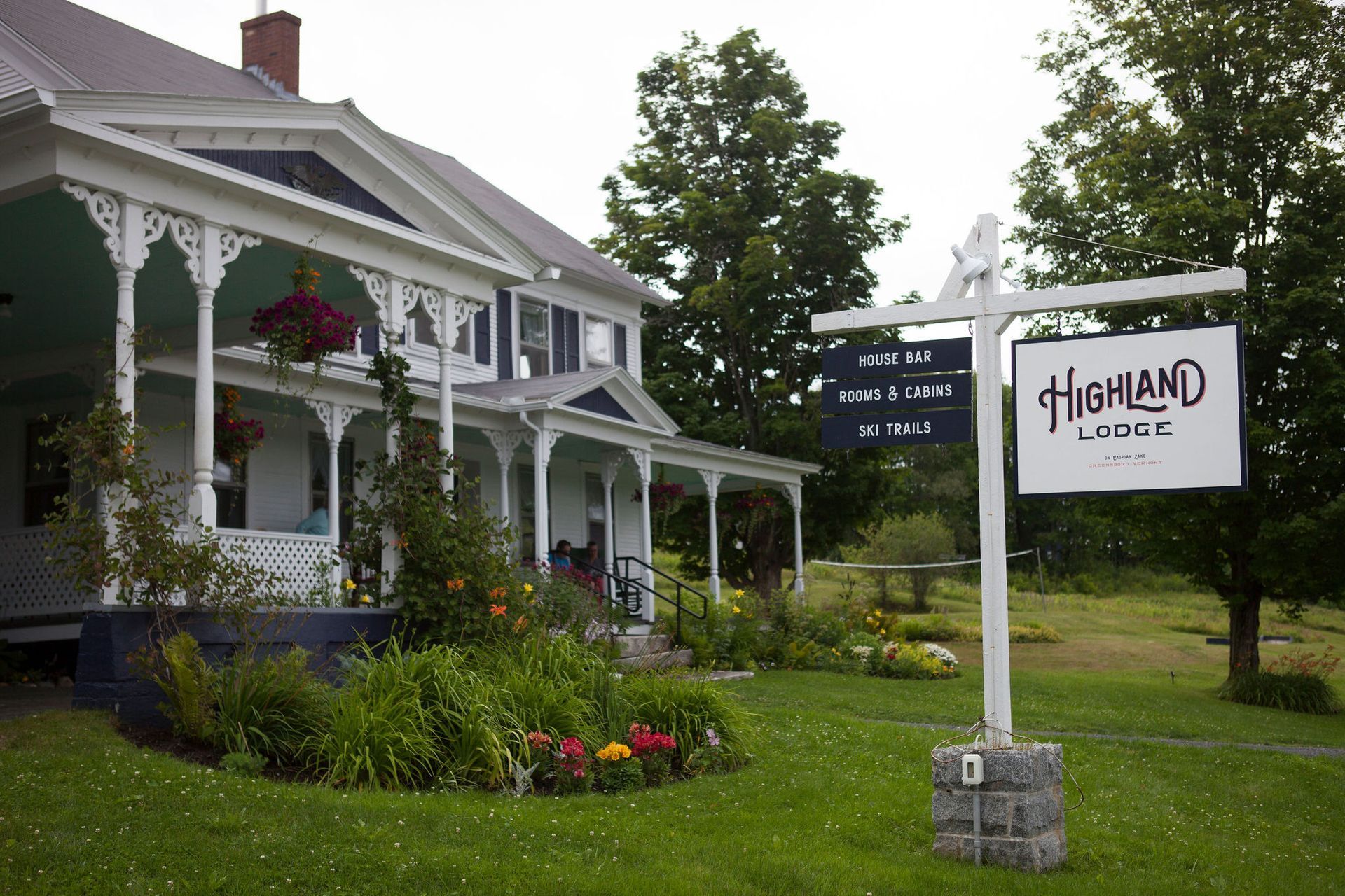Highland Lodge with white porch and sign on a green lawn, flowers, overcast sky.