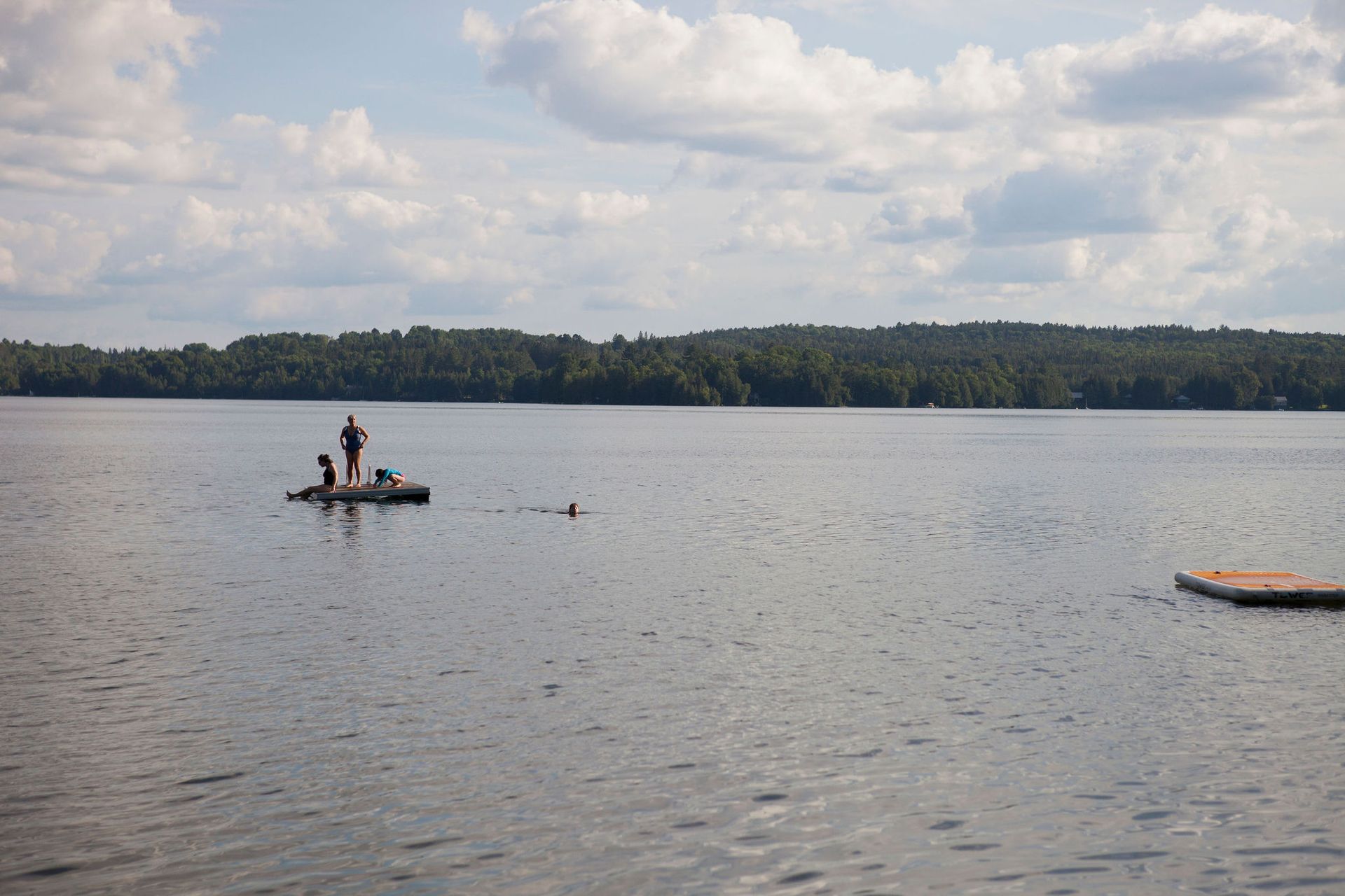 Lake scene with a small raft and swimmers. Forested shoreline under a cloudy sky.