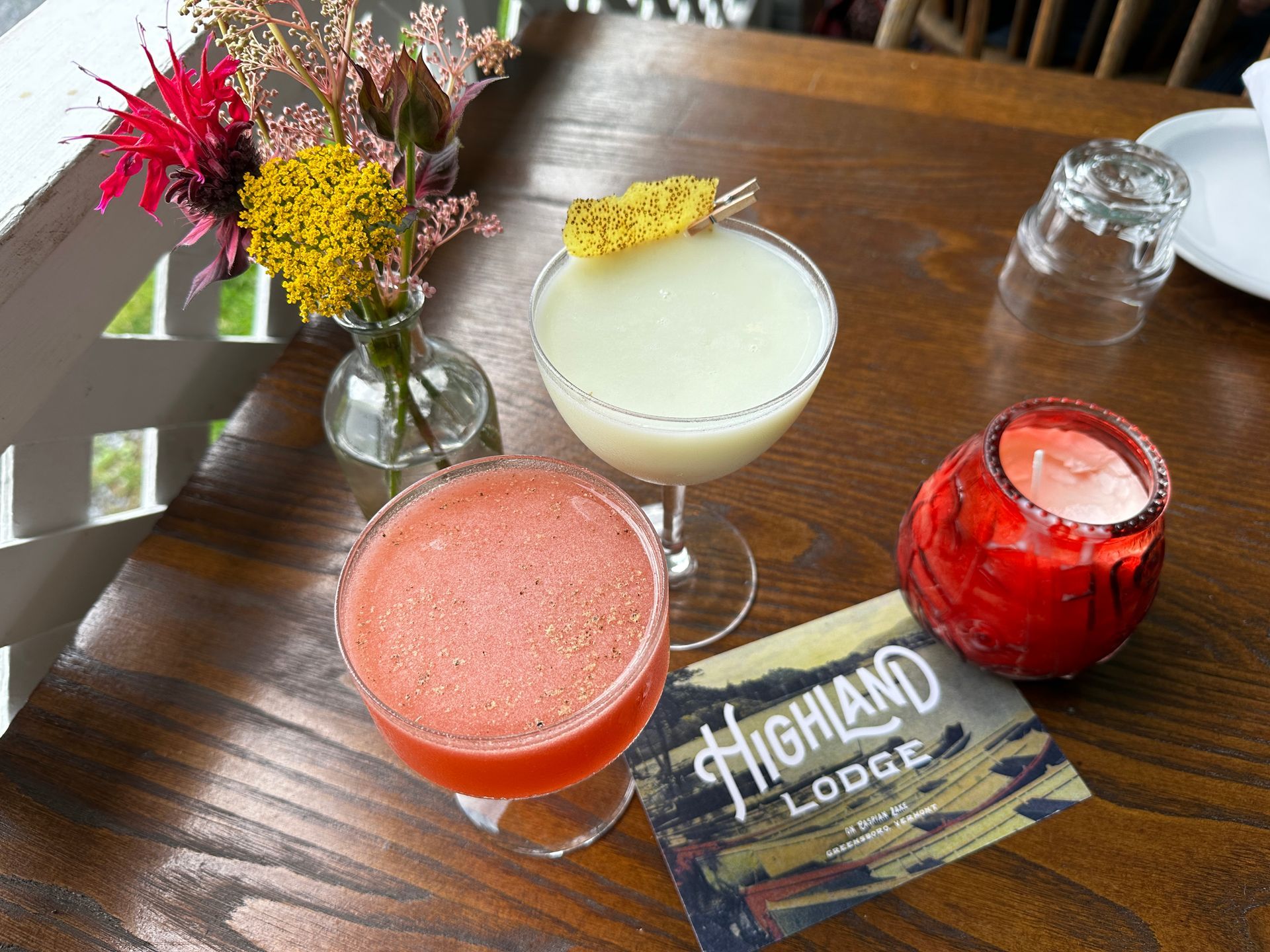 Two cocktails, a red one and a white one, on a wooden table with a card and flowers.