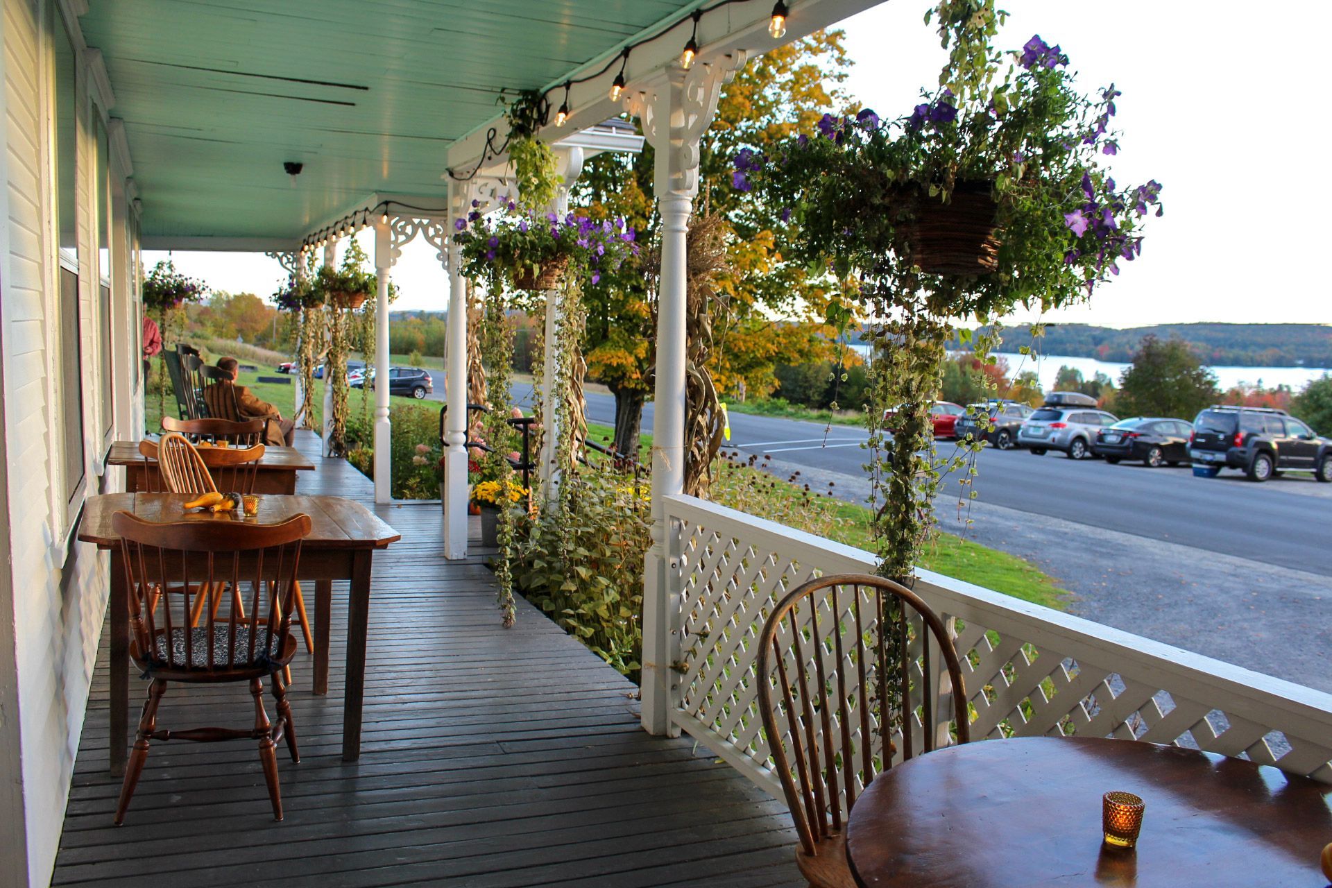 Outdoor restaurant patio with tables, chairs, and hanging flowers overlooking a road and water.