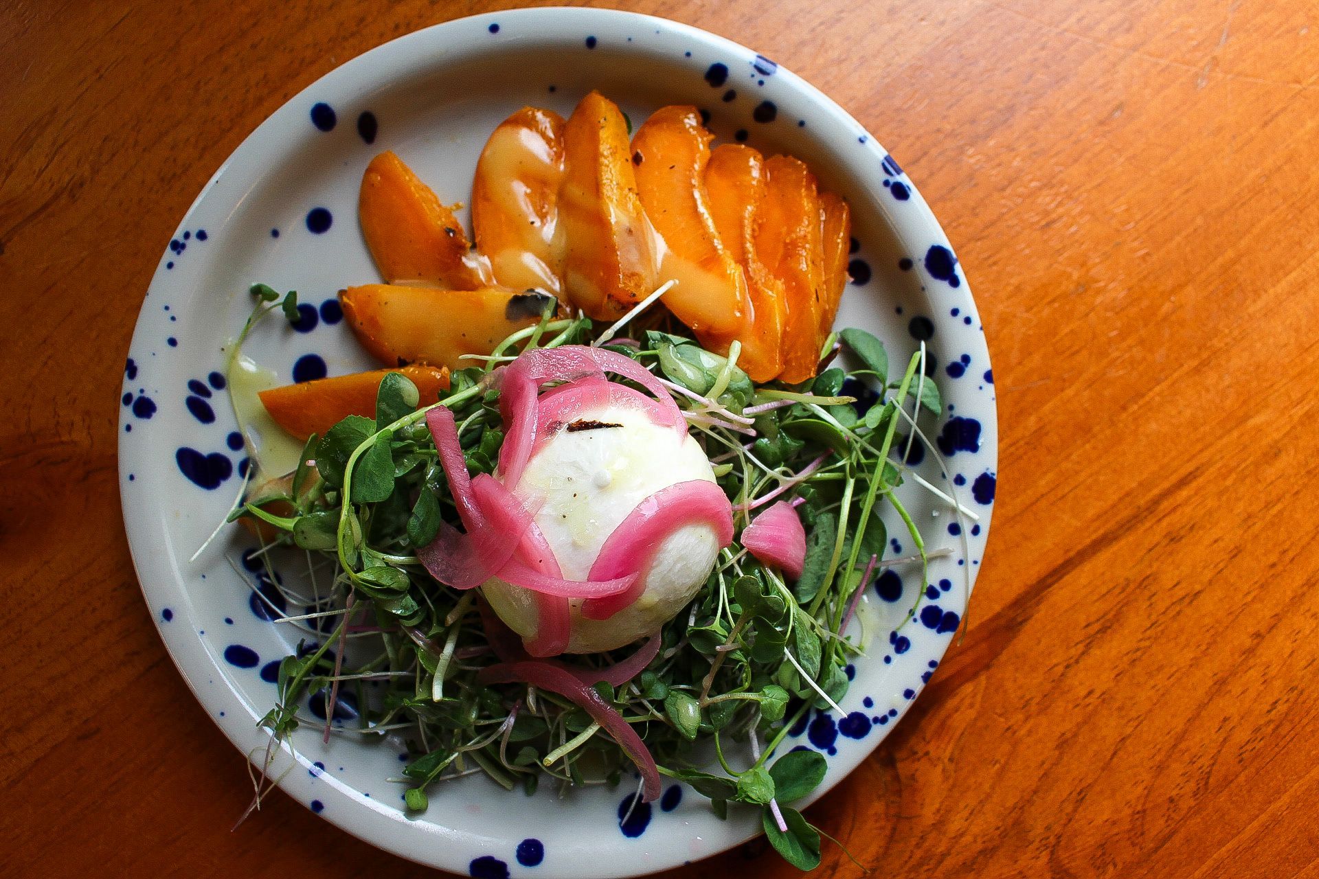 Plate with peaches, greens, a white ball topped with pink onions, on a wood table.