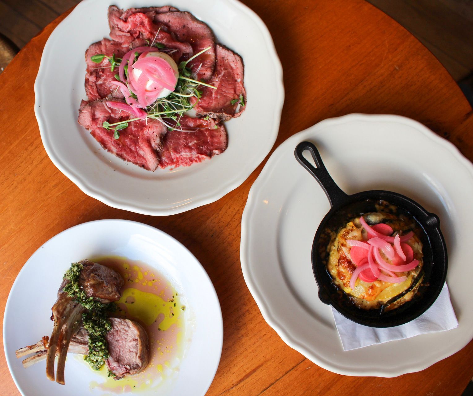 Three plates of food on a table: sliced meat with pink onion, a dish in a cast iron pan, and lamb chops.