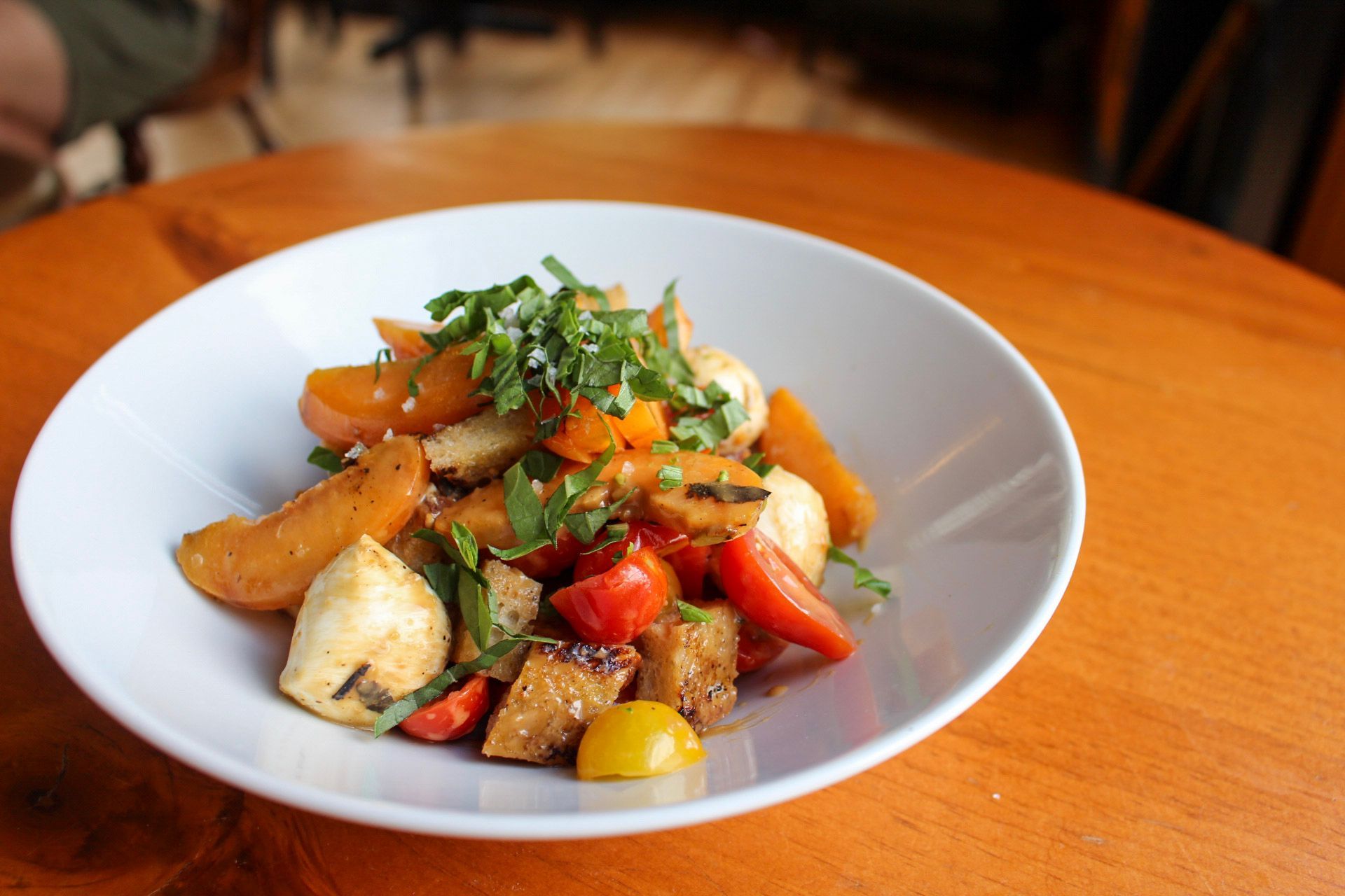 Salad with mozzarella balls, apricots, and tomatoes in a white bowl on a wooden table.
