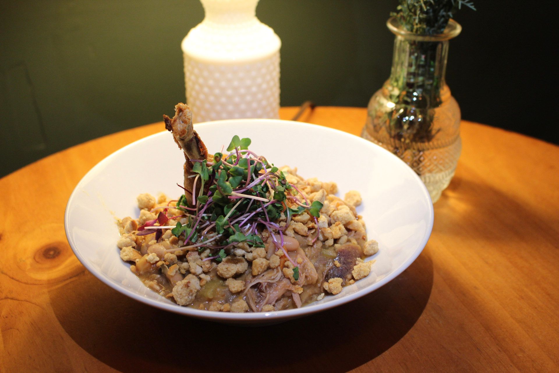 Bowl of food on a wooden table, topped with sprouts and a fried item. A bottle and vase are in the background.
