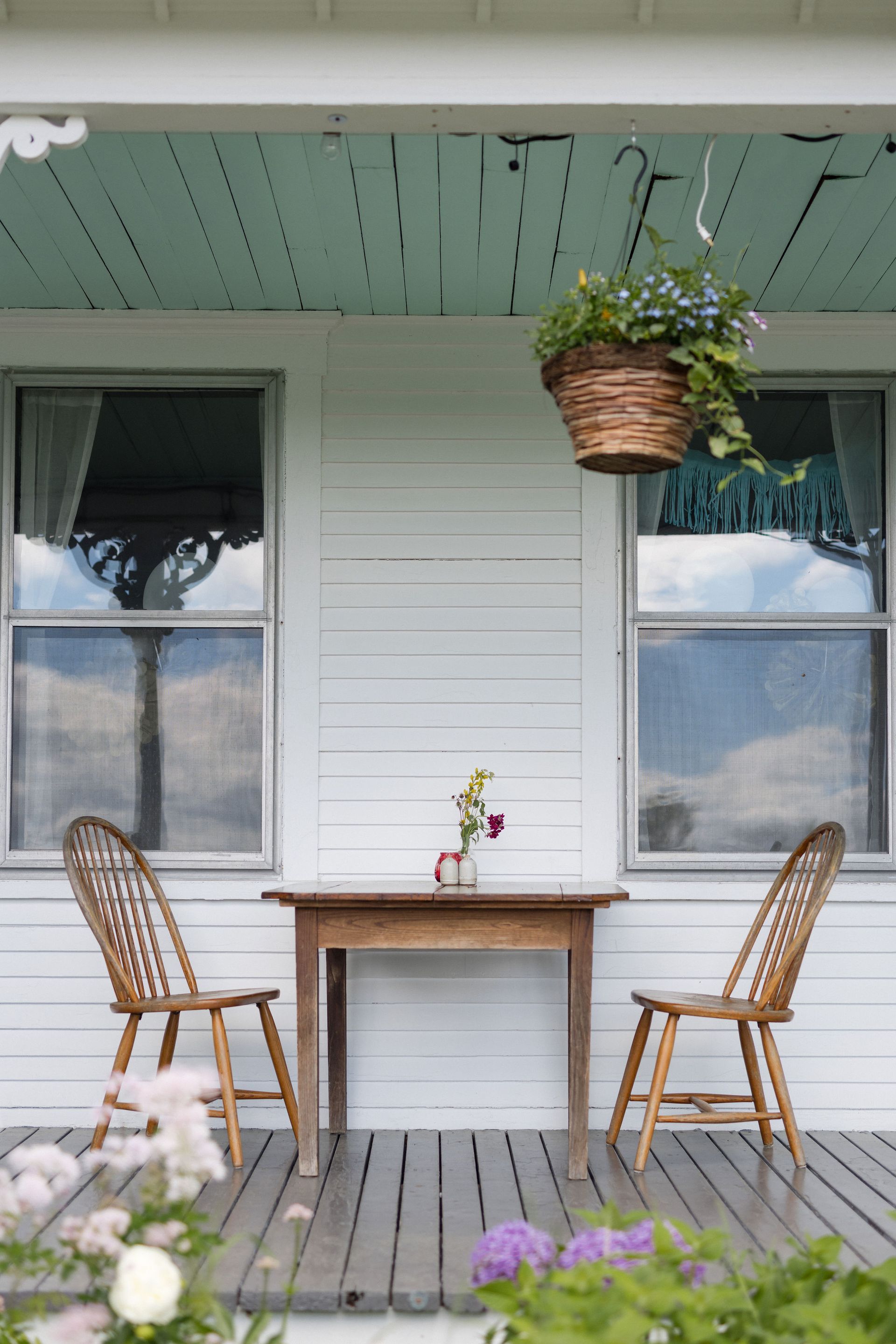 White porch with wooden table, chairs, and hanging basket with flowers.