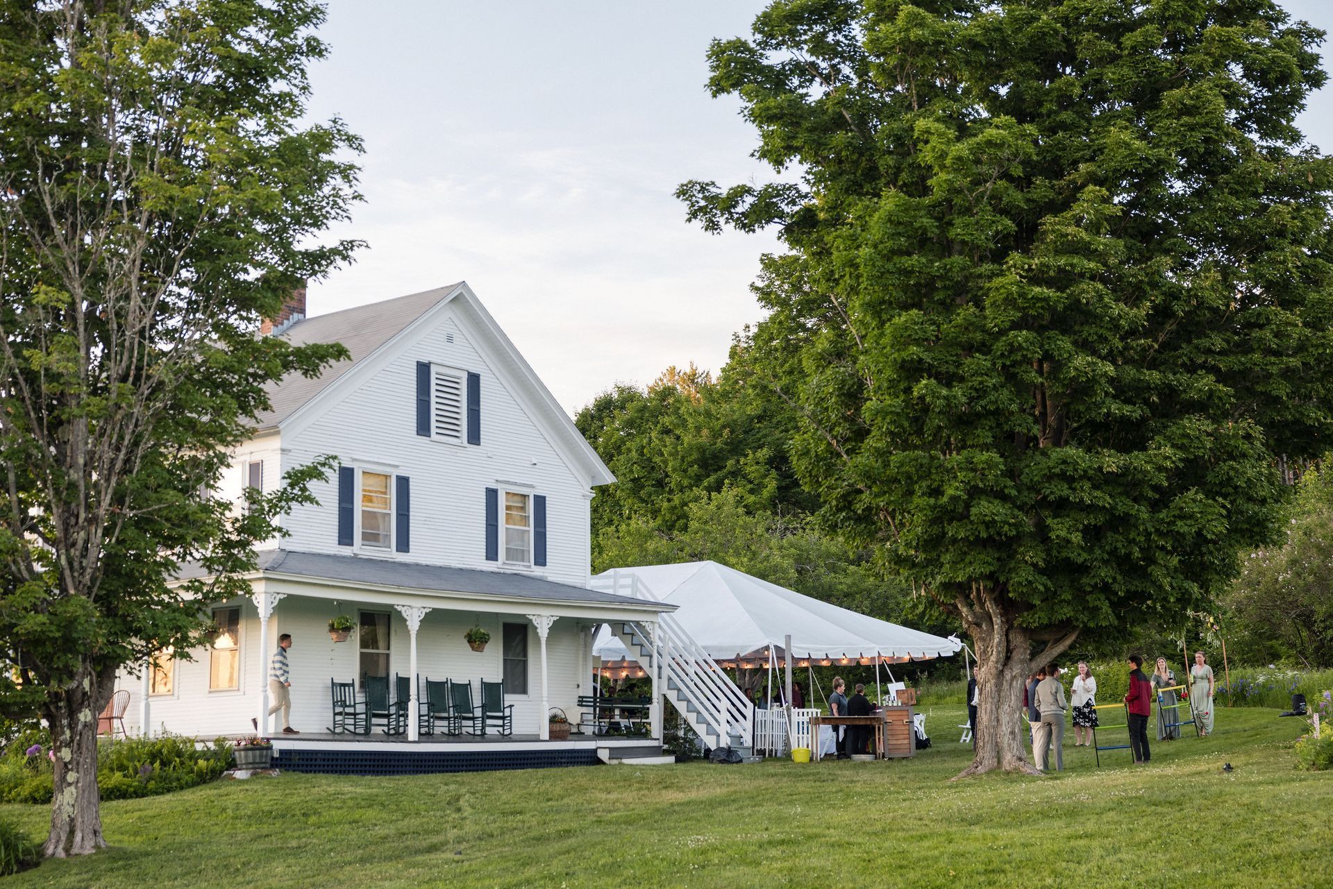 White house with porch, tent for event, people gathered on a lawn, green trees, evening light.
