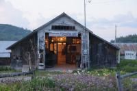 Old wooden barn entrance, doorway glowing with interior lights, surrounded by greenery.