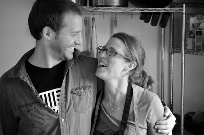 Man and woman smiling, embracing. Kitchen setting with shelves and utensils visible.