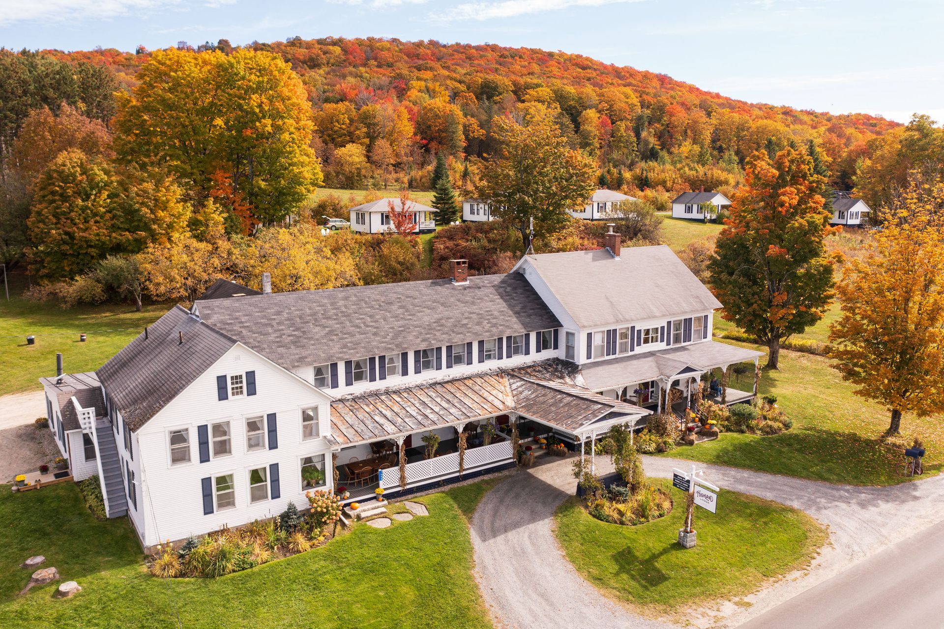 White building with a porch, autumn trees with colorful leaves, and a hillside in the background.