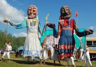 Two large puppets are held up by people in a field. Blue sky, circus backdrop.