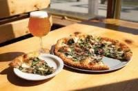 Pizza slice on a small plate next to a whole pizza on a metal pan and a glass of beer, on a wooden surface near a window.