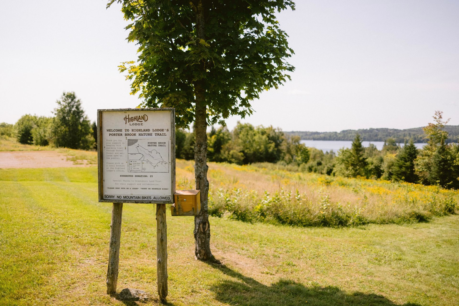 Sign with text and map stands in a grassy field with a tree. Distant trees and a lake in the background.