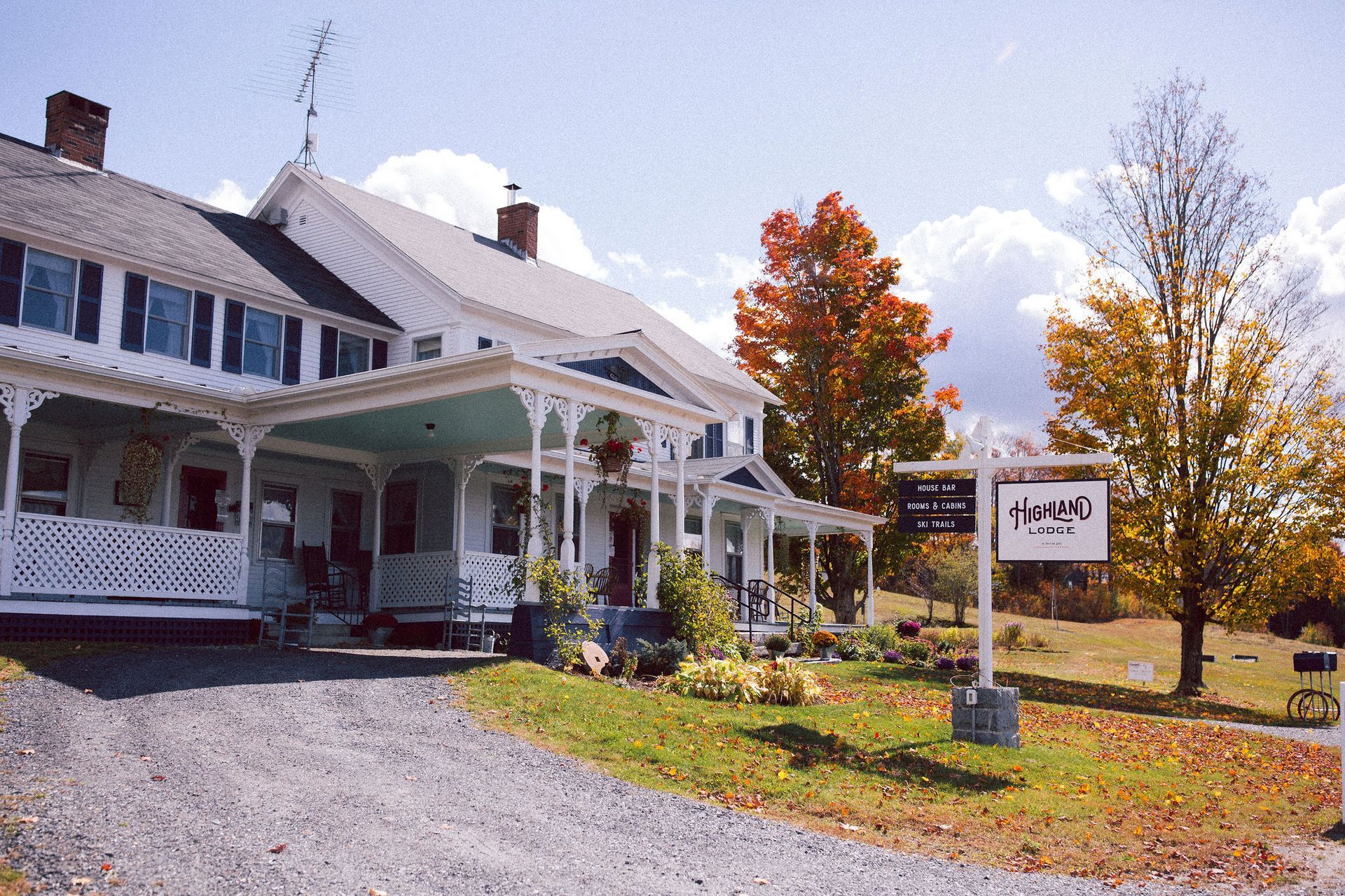 A white Victorian inn with a porch, a gravel driveway, and a sign against an autumn landscape.