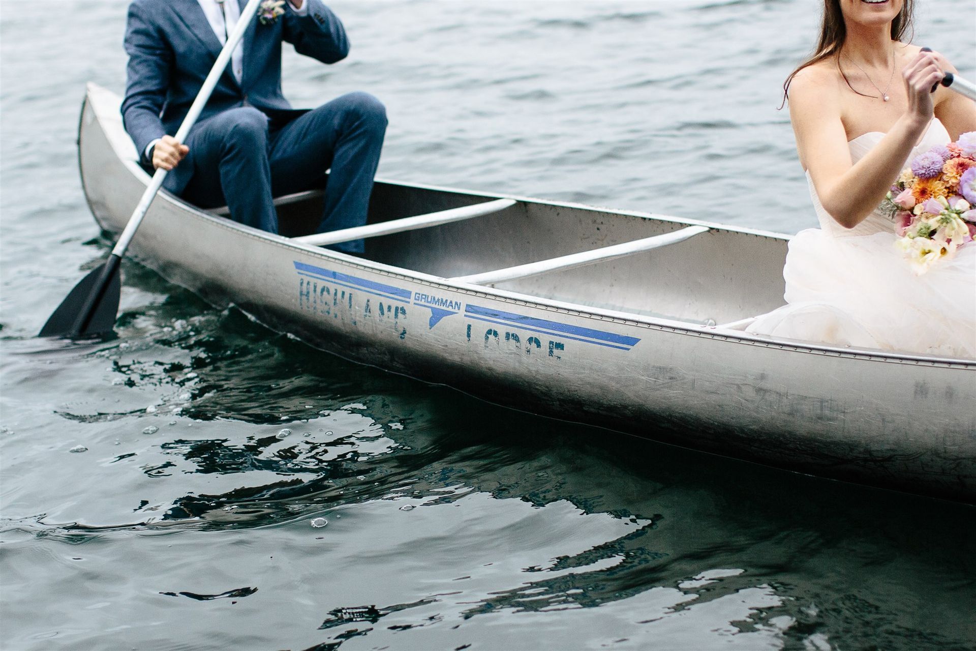 Bride and groom in a canoe on water; the man paddles, the woman holds flowers.