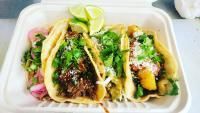 Three tacos in a container, topped with cilantro, onions, lime, and cheese.
