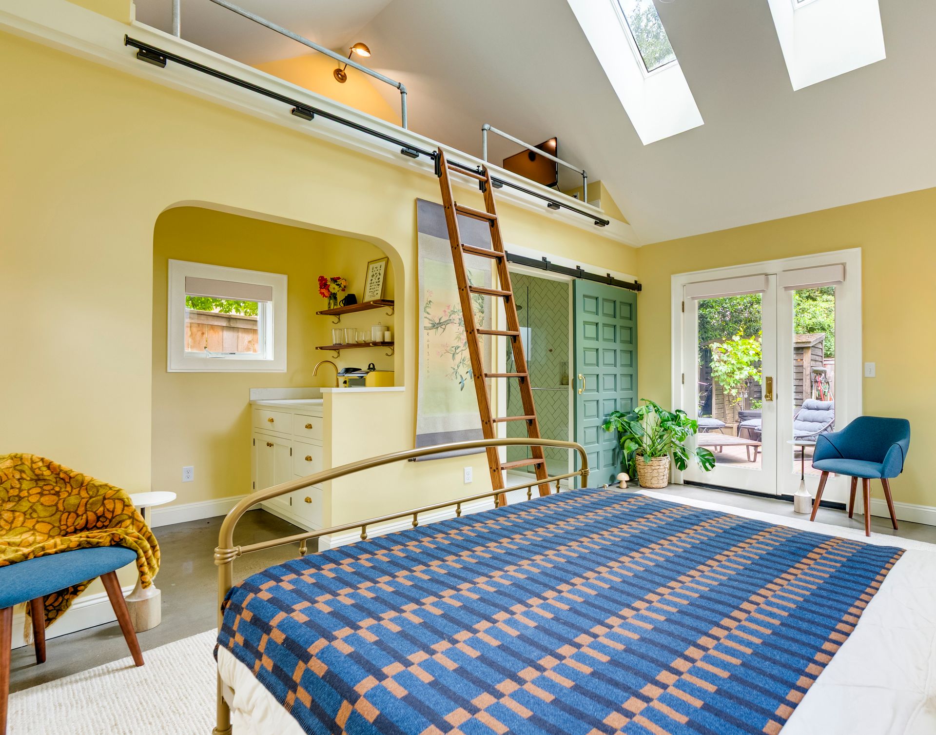 A cheery butter yellow garage-turned-studio with a loft above a kitchenette and cozy bed in the foreground.