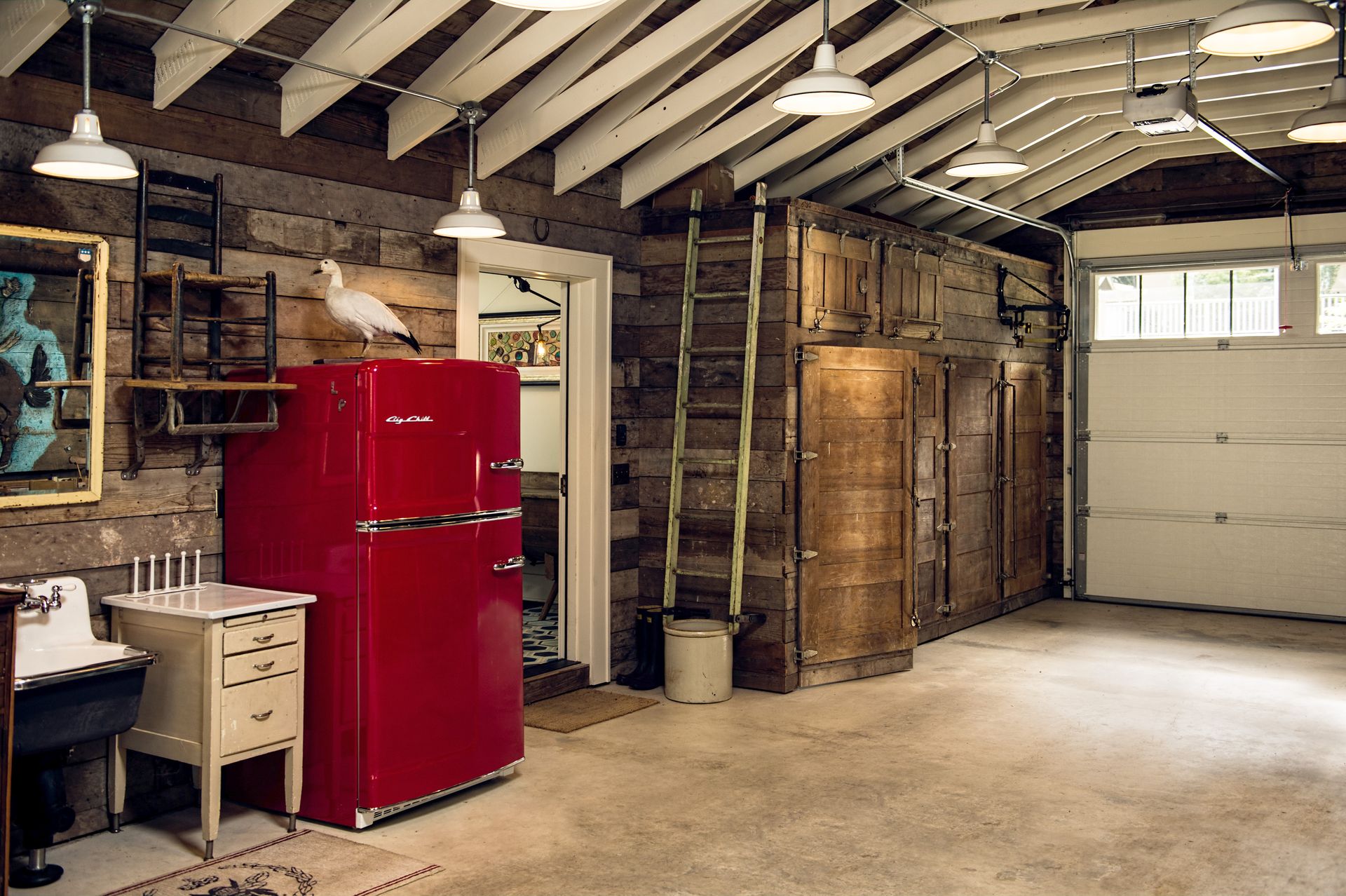 Salvaged wood paneling contrasts with white scissor trusses in the garage. A retro red fridge and custom built in cabinet flank the door to the mudroom.
