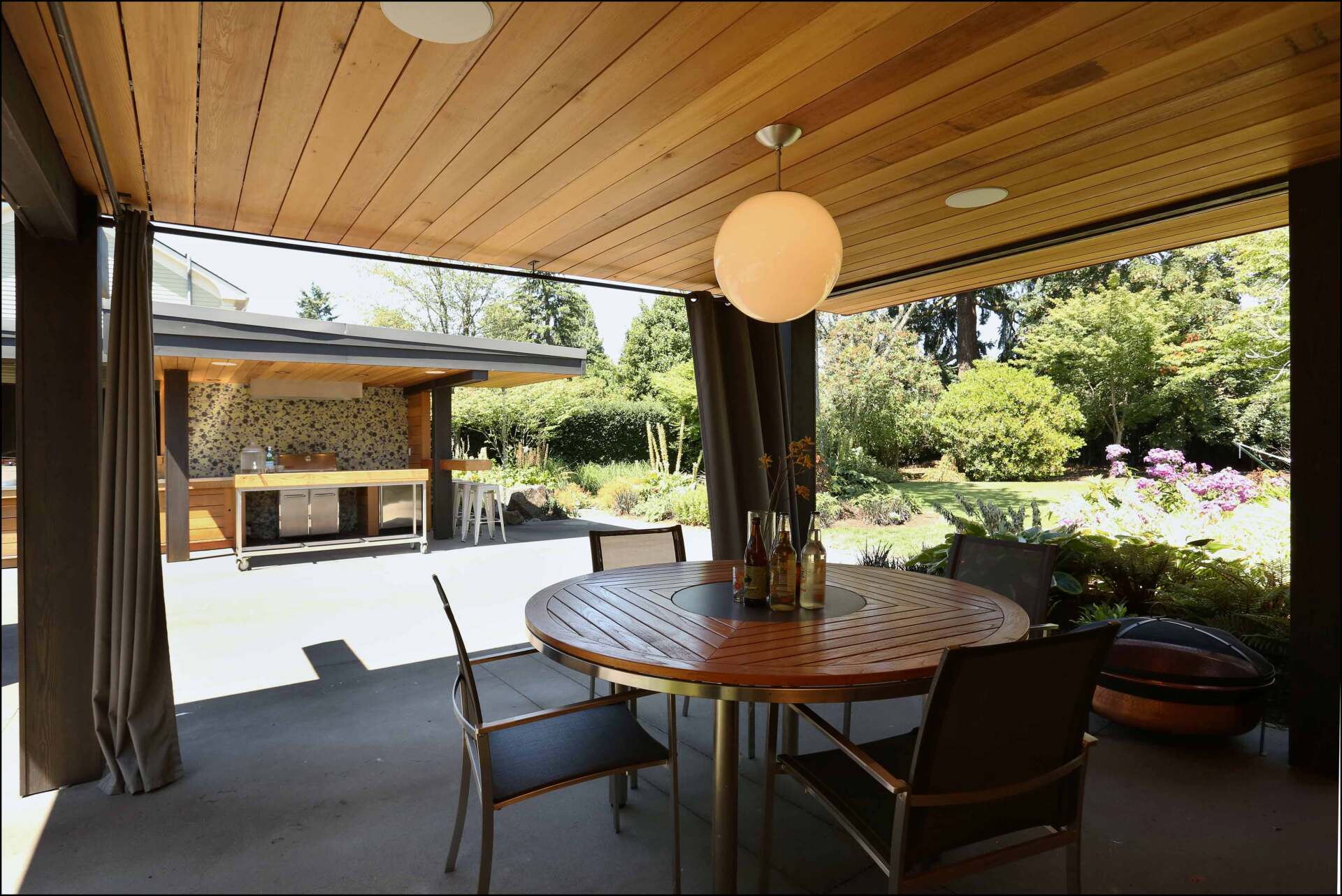 View of the outdoor kitchen from a generous dining pavilion with round table, wood paneled ceiling and globe pendant light.