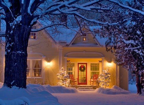 Snowy house with warm porch lights glowing at dusk, framed by snow-covered trees.