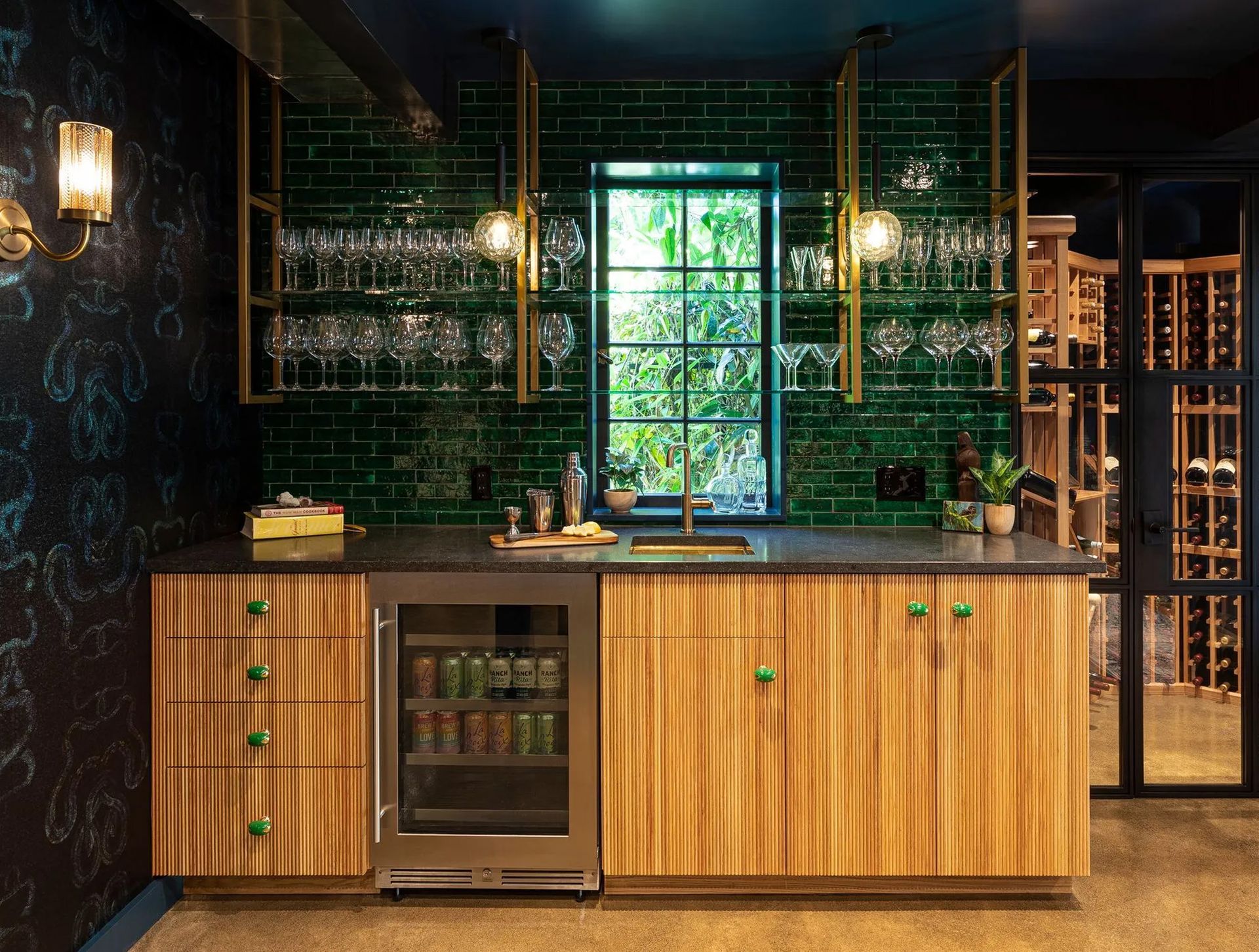 Against the dark backdrop, glossy green tile, brass, glass, and warm wood reflect and amplify the available light, giving the basement bar a rich sense of depth and warmth.