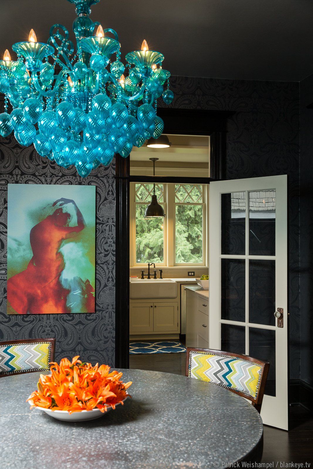 The dining room has black tone-on-tone wallpaper, dark floors, colorful artwork and a turquoise glass chandelier as well as natural light from the windows. The kitchen is visible through the open door beyond.