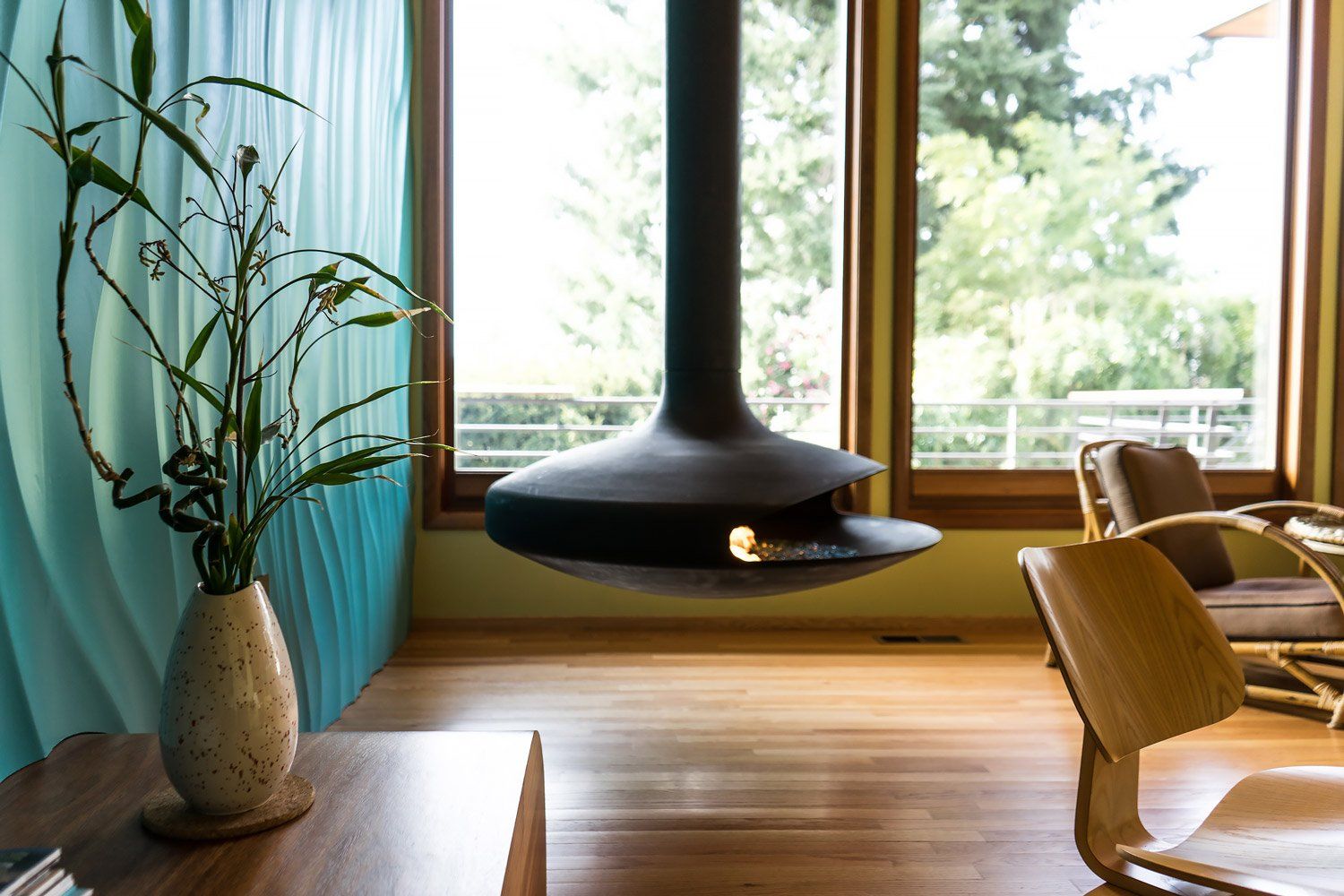 In the living room, a corner gyro focus fireplace hangs from the ceiling with the textured wall and full height windows behind it.