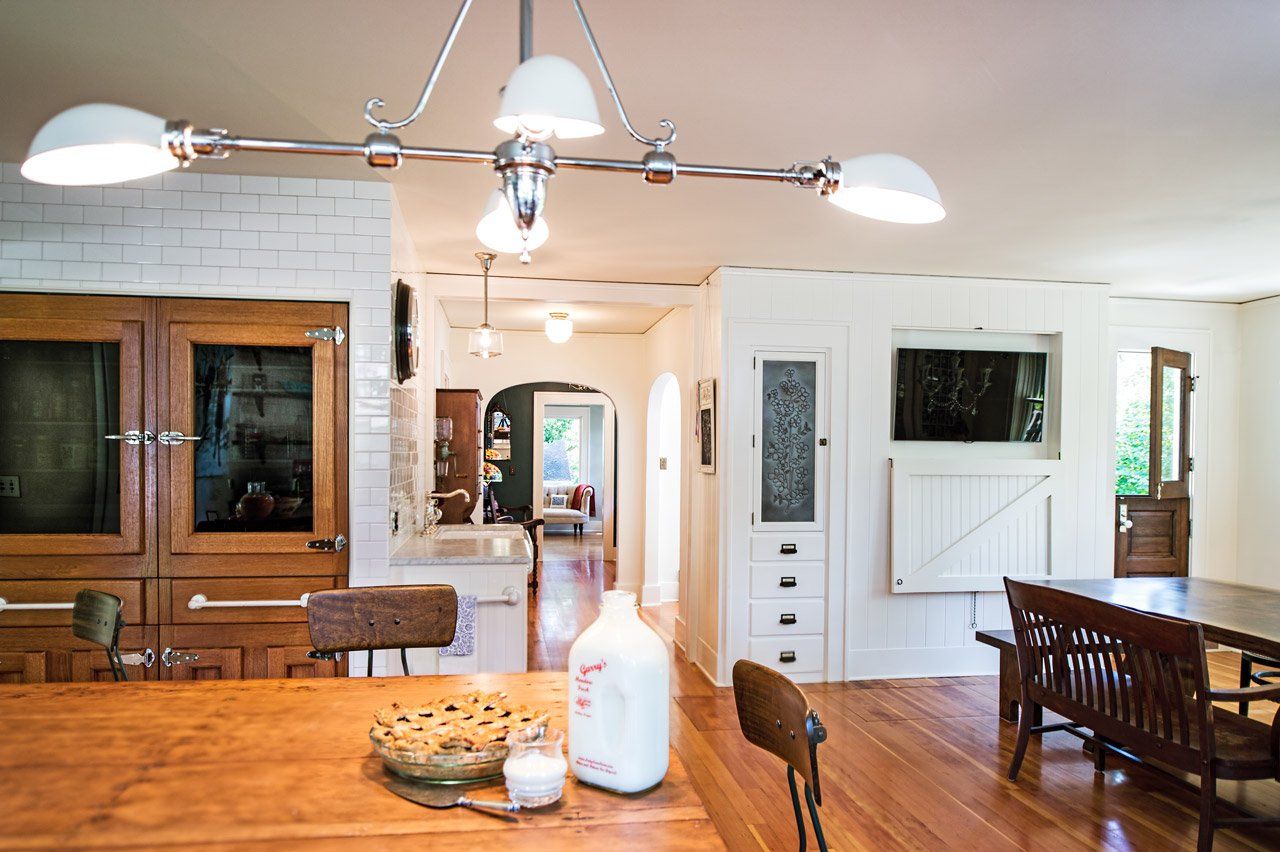 Wide view of the kitchen over an antique table, with customized fridge with wooden doors, a dutch door to the side yard and the hall to the living room in between.
