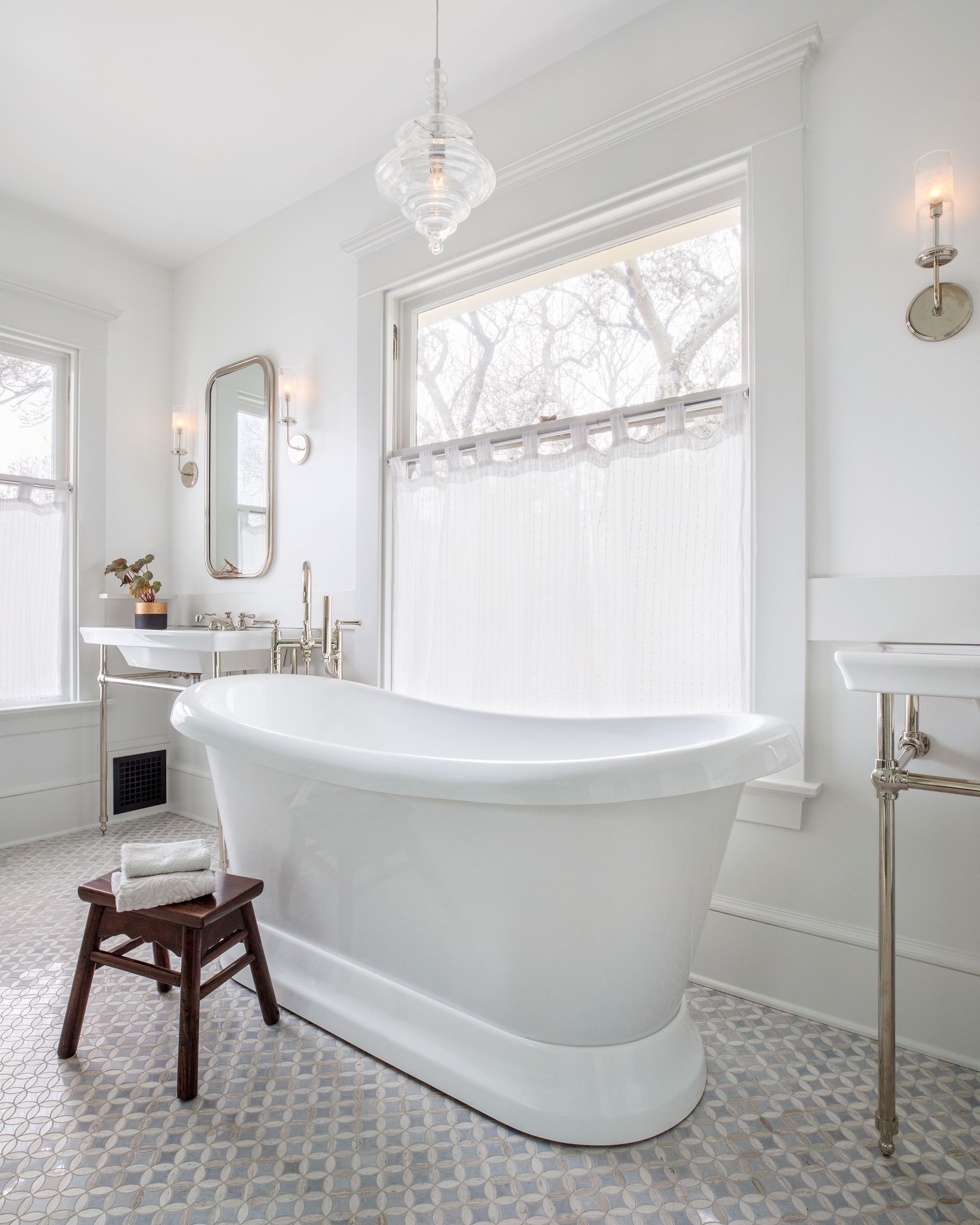 An airy bathroom, formerly a bedroom, features console sinks, a freestanding tub and marble mosaic floors. 