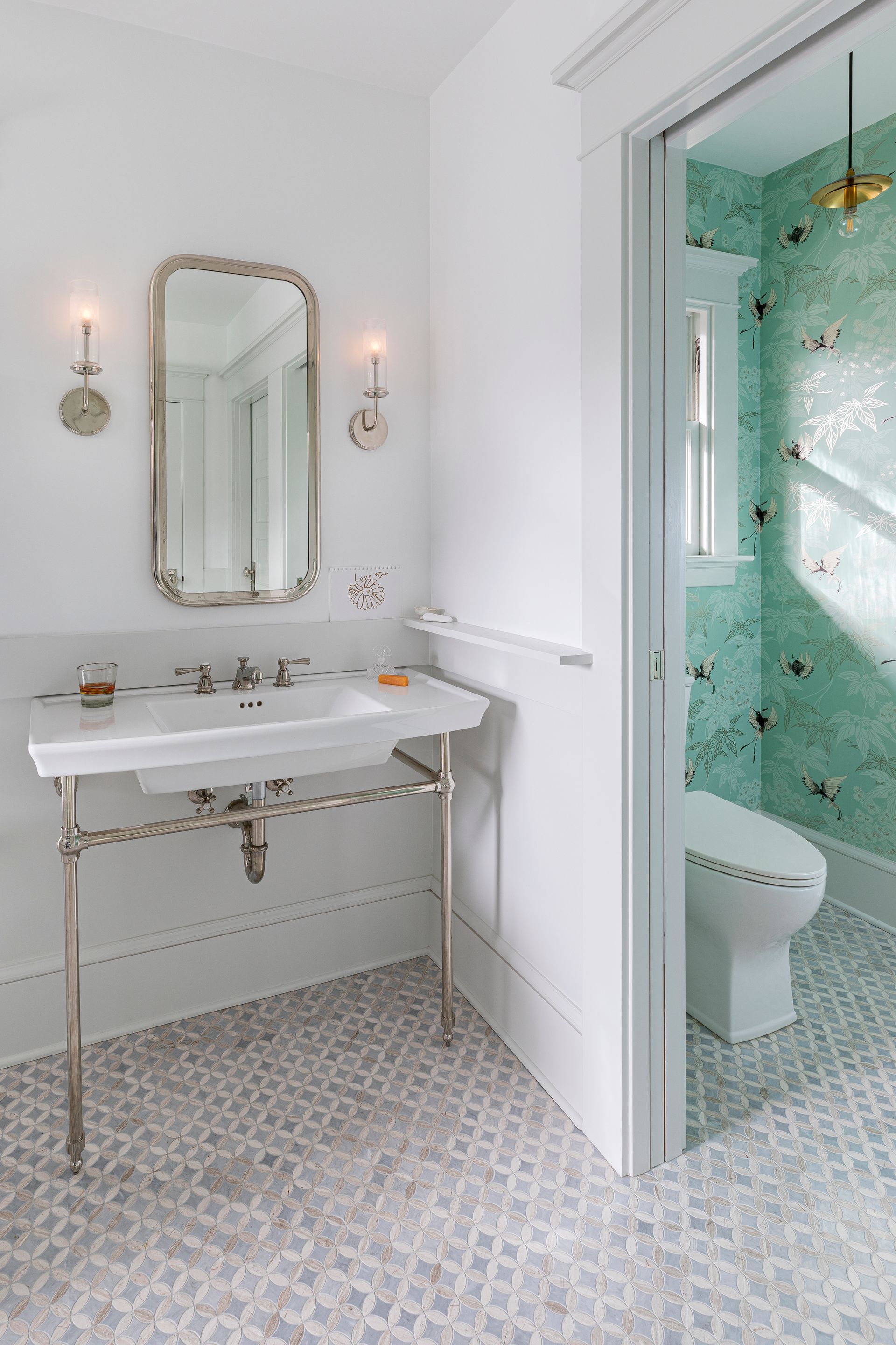 A airy bathroom with a console sink and mosaic stone floor features a private WC with hummingbird wallpaper.